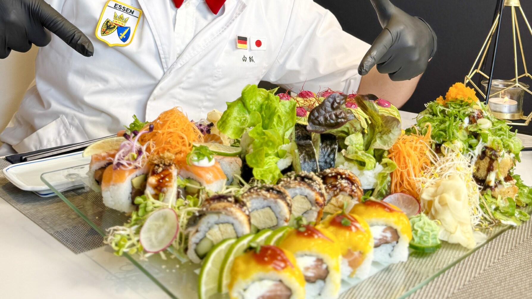 Auf einem Tisch steht ein Teller mit Sushi, im Hintergrund sitzt eine Person und Hände zeigen auf den Teller.