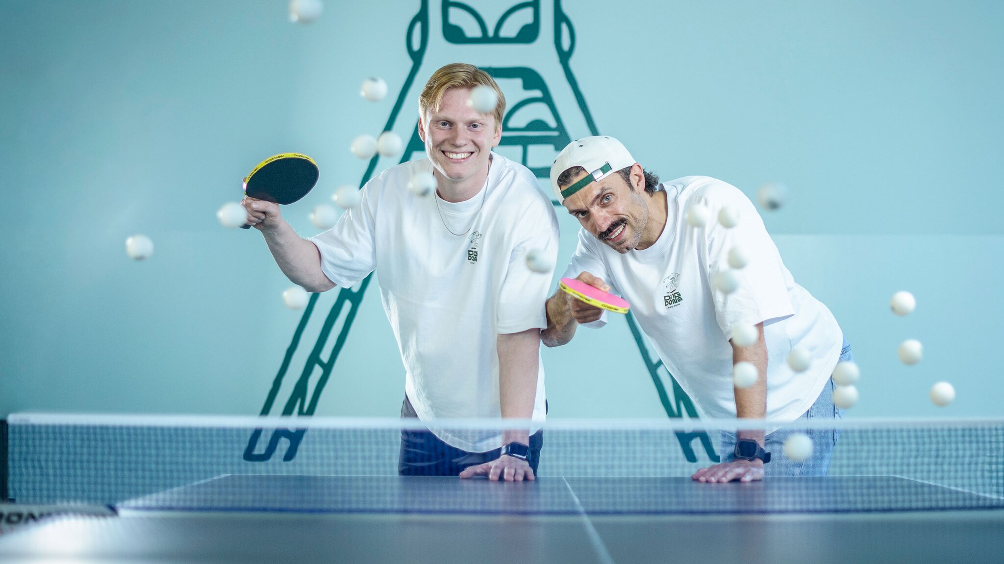 Zwei der Kamera zugewandte lächelnde Personen mit Tischtennisschlägern in der Hand an einer Tischtennisplatte, um die herum einige Tischtennisbälle fliegen.