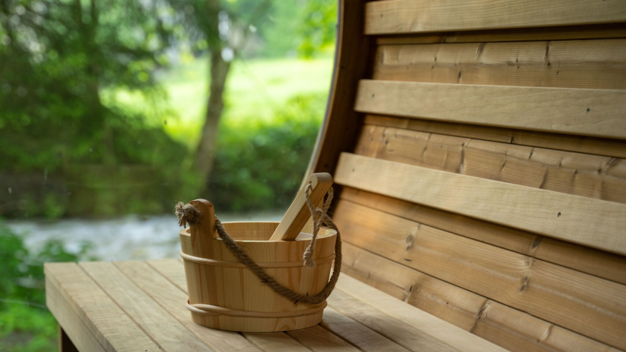 Ein Holzgefäß mit Kelle steht auf einer hölzernen Sitzbank mit abgerundeter Rückenfläche im Grünen.