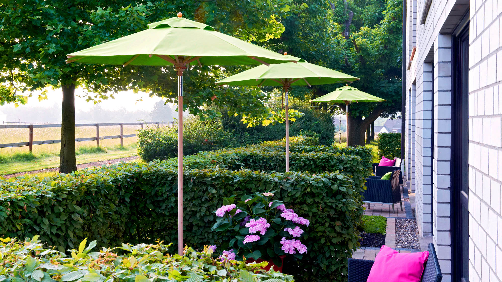 Im Außenbereich eines Hotels sind kleine, private Terrassen mit Blick auf eine Wiese. Jede ist mit Sonnenschirm ausgestattet und einer Hecke als Sichtschutz.