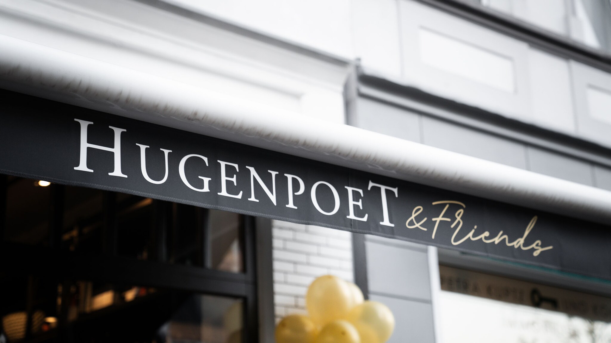 Eine weiße Gebäudefassade mit dunklem Fenster und goldenen Ballons, über der eine Markise ausgefahren ist, auf der der Schriftzug „Hugenpoet & Friends“ steht.