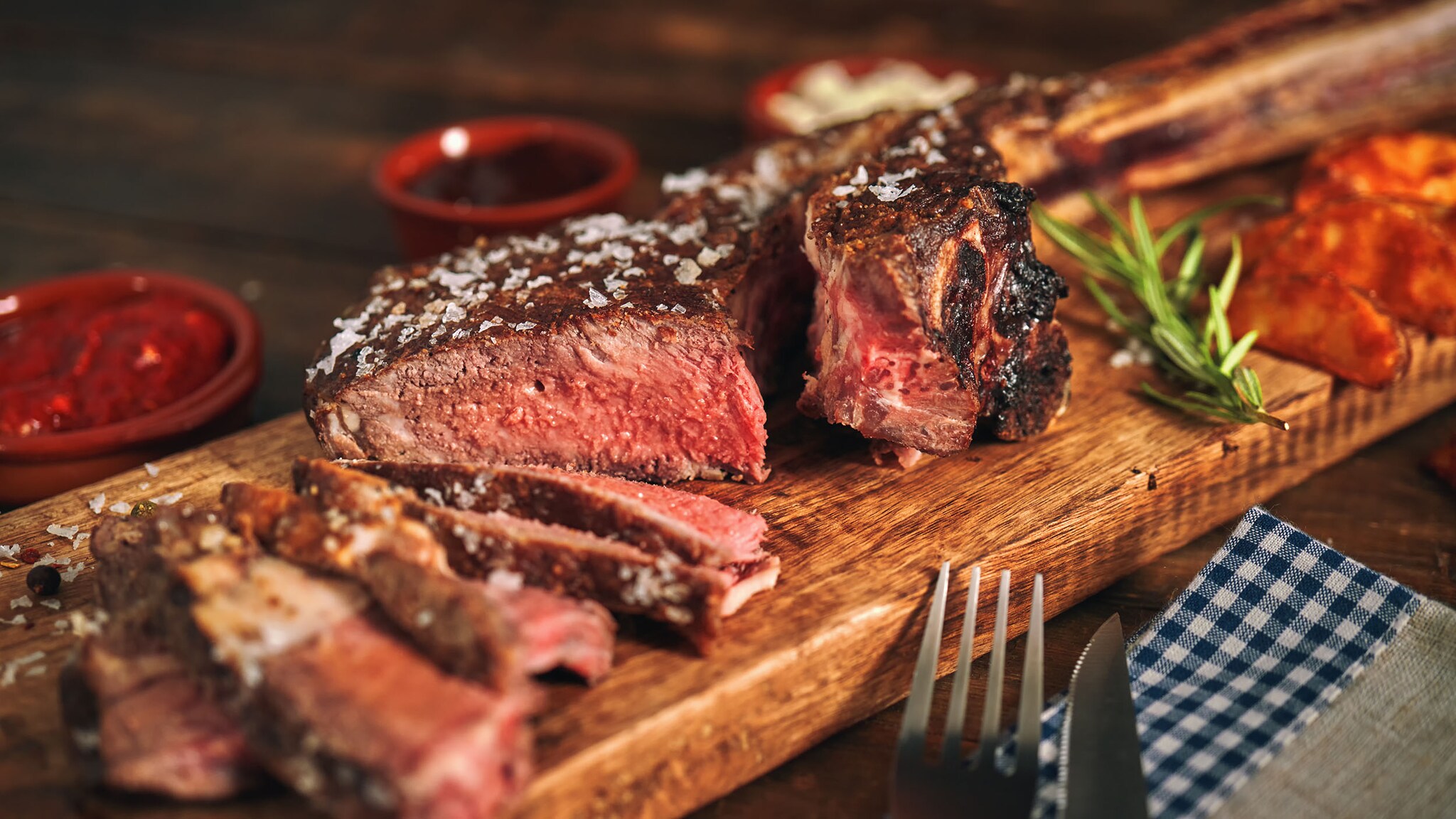 Ein saftiges Steak, das in mehrere Scheiben und Stücke geschnitten wurde, liegt auf einem Holzbrett, daneben Gabel und Messer auf einer Serviette. 