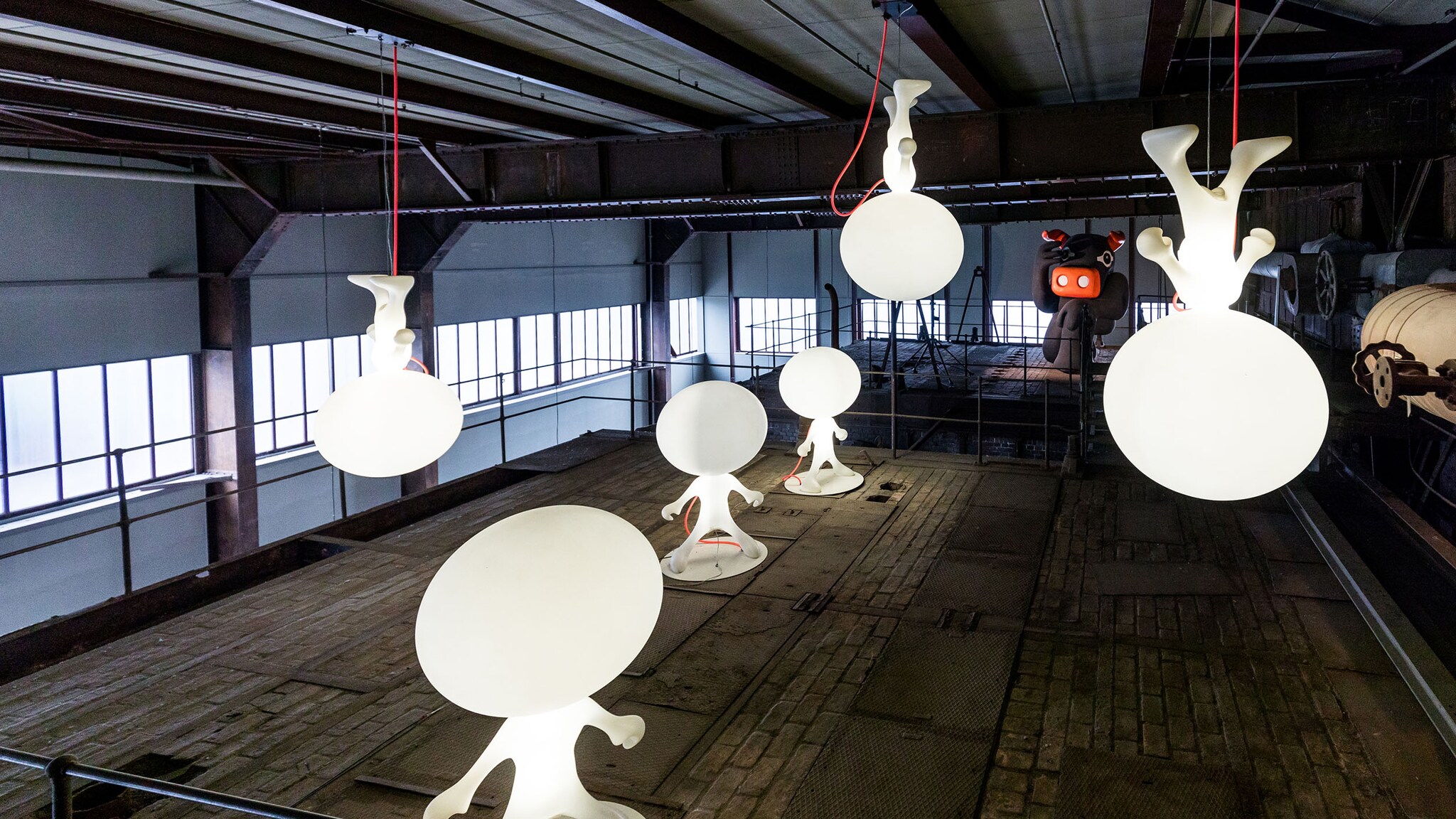 Mehrere leuchtende Skulpturen in Form von Männchen mit großem Kopf in einer großen Lagerhalle, die als Museum dient.