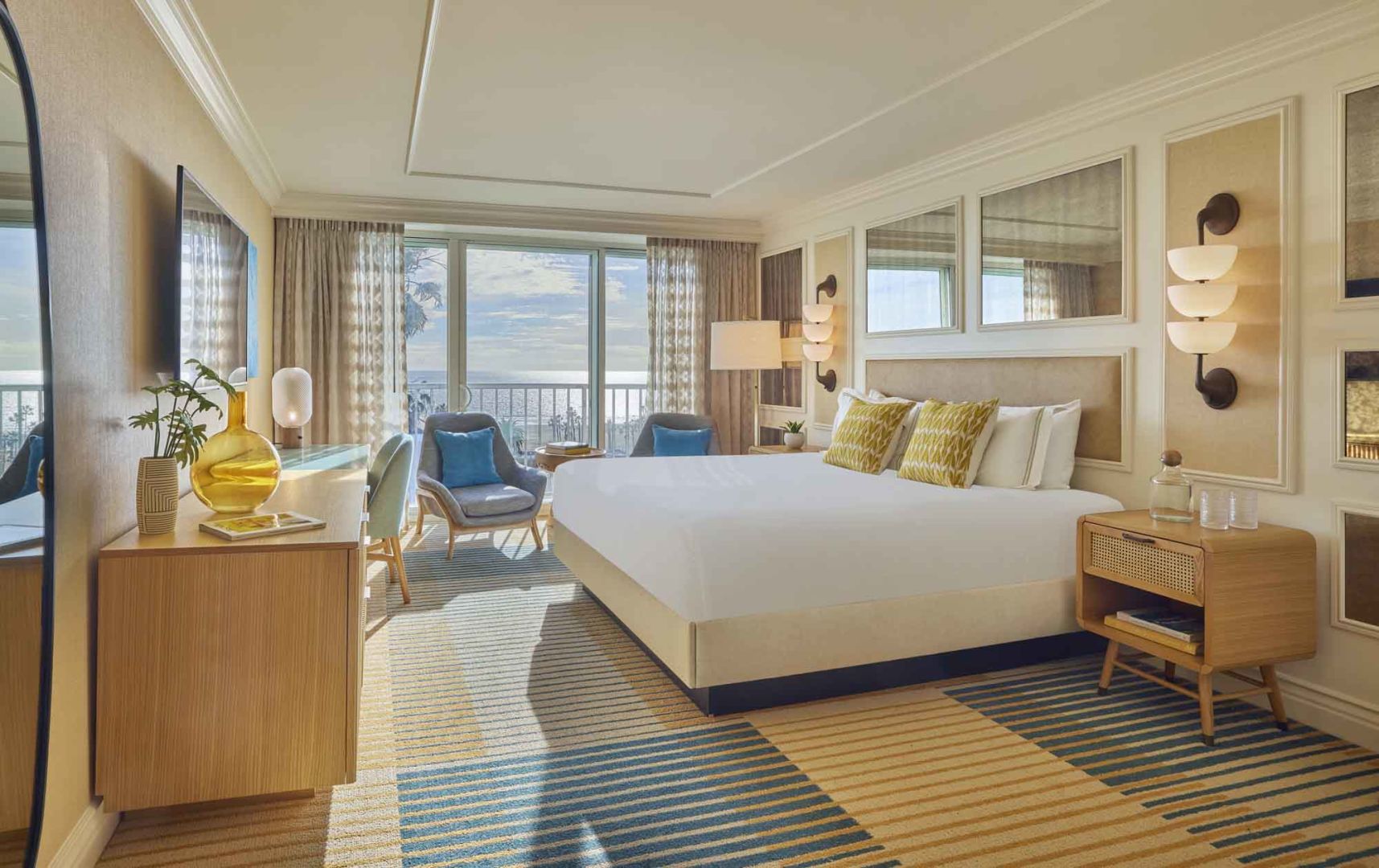 Ein sonnendurchflutetes Hotelzimmer mit Doppelbett und Meerblick in cremefarbenen Tönen und Akzenten in türkis, beige und goldgelb.