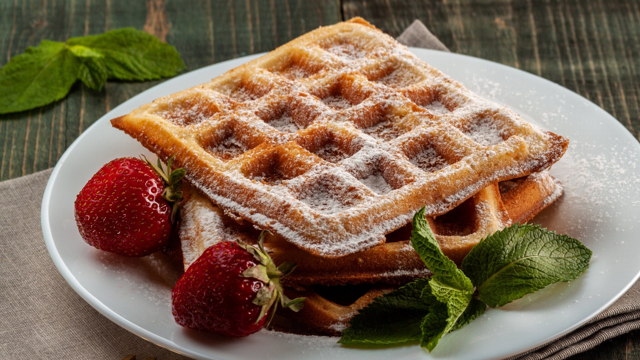 Ein weißer Teller mit einem Stapel belgischer Waffeln, die mit Puderzucker bedeckt sind, daneben Minzblätter und zwei Erdbeeren.
