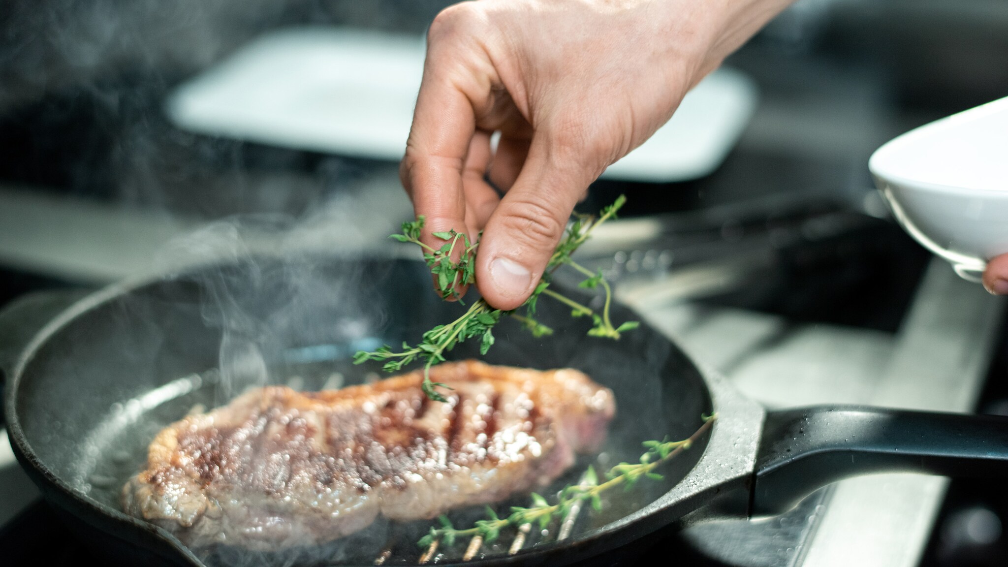 Ein saftiges Steak, das in einer Pfanne gebraten und mit Thymian verfeinert wird, darüber eine Hand mit mehreren Zweigen. 