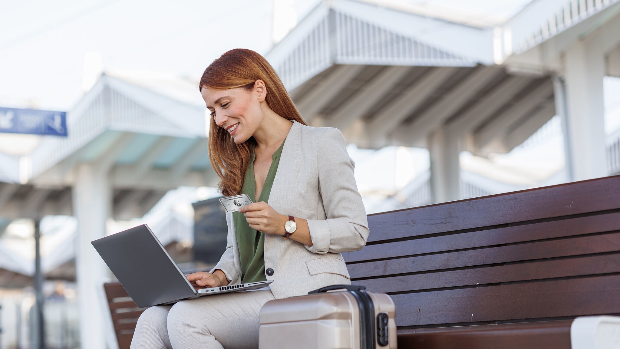Eine Frau sitzt auf einer Bank an einem Bahnhof, hat ihren Laptop auf dem Schoß und eine Kreditkarte in der Hand.