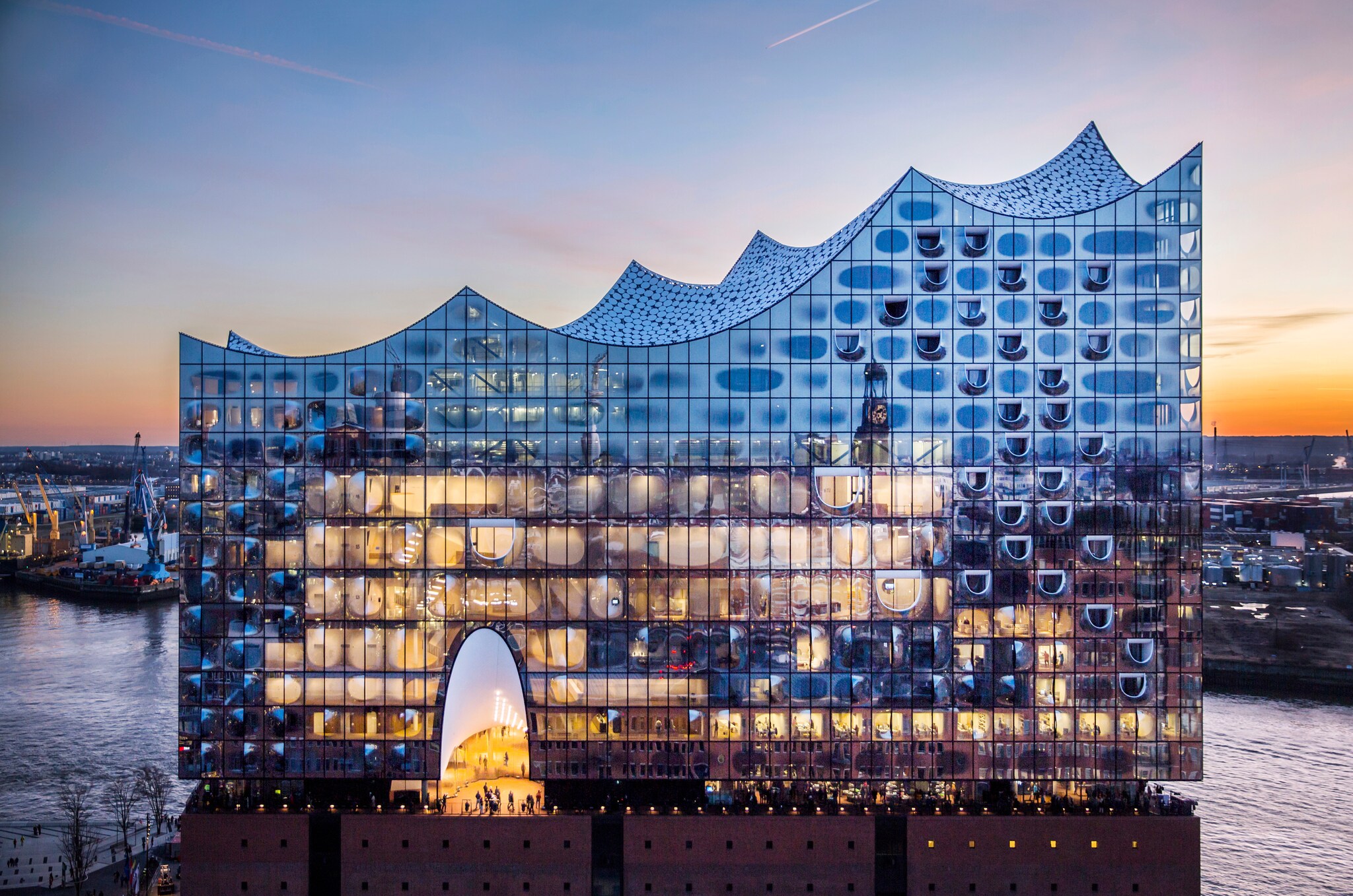Die Elbphilharmonie in Hamburg bei Sonnenuntergang: Die gläserne, wellenförmige Fassade leuchtet über dem Hafen, während sich der Himmel rosa und blau färbt.