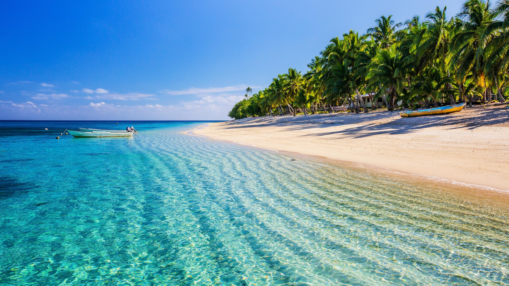 Sandstrand mit Palmen und Booten am türkisblauen Meer, auf dem ein Boot treibt.
