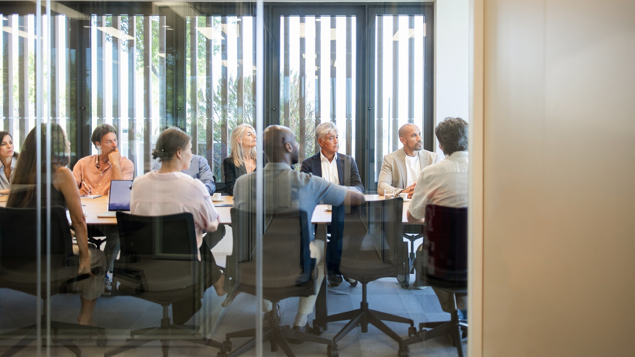 Büroraum hinter einer Glasscheibe an einem langen Tisch mit vielen Bürostühlen, auf denen verschiedene Personen sitzen und den Blick in eine Richtung gewandt haben.