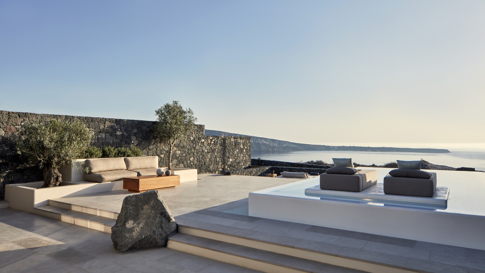 Private Steinterrasse eines Hotelzimmers mit Sofas, einem Infinitypool und Liegen mit Blick aufs Meer.