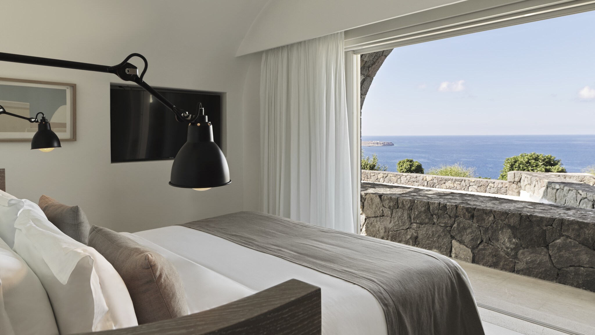 Ein Hotelzimmer mit Bett, das zu einer großen Fensterfront ausgerichtet ist, im Hintergrund Steinmauern und Meer.