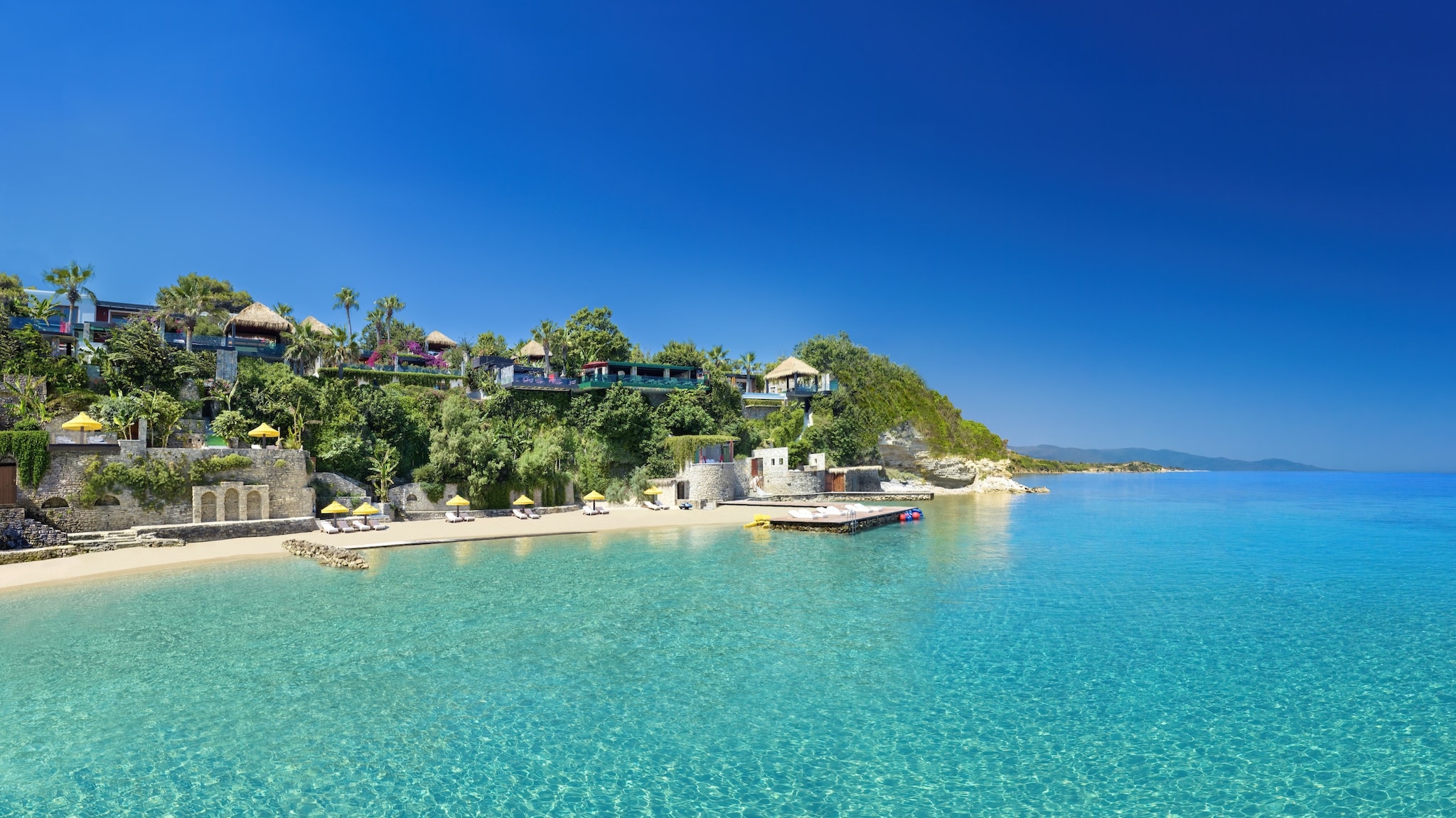 Porto Zante, Sandy Beach Blauer Himmel über türkisblauem Meer. An der Küste sind ein Sandstrand, Palmen und kleine Gebäude erkennbar.
