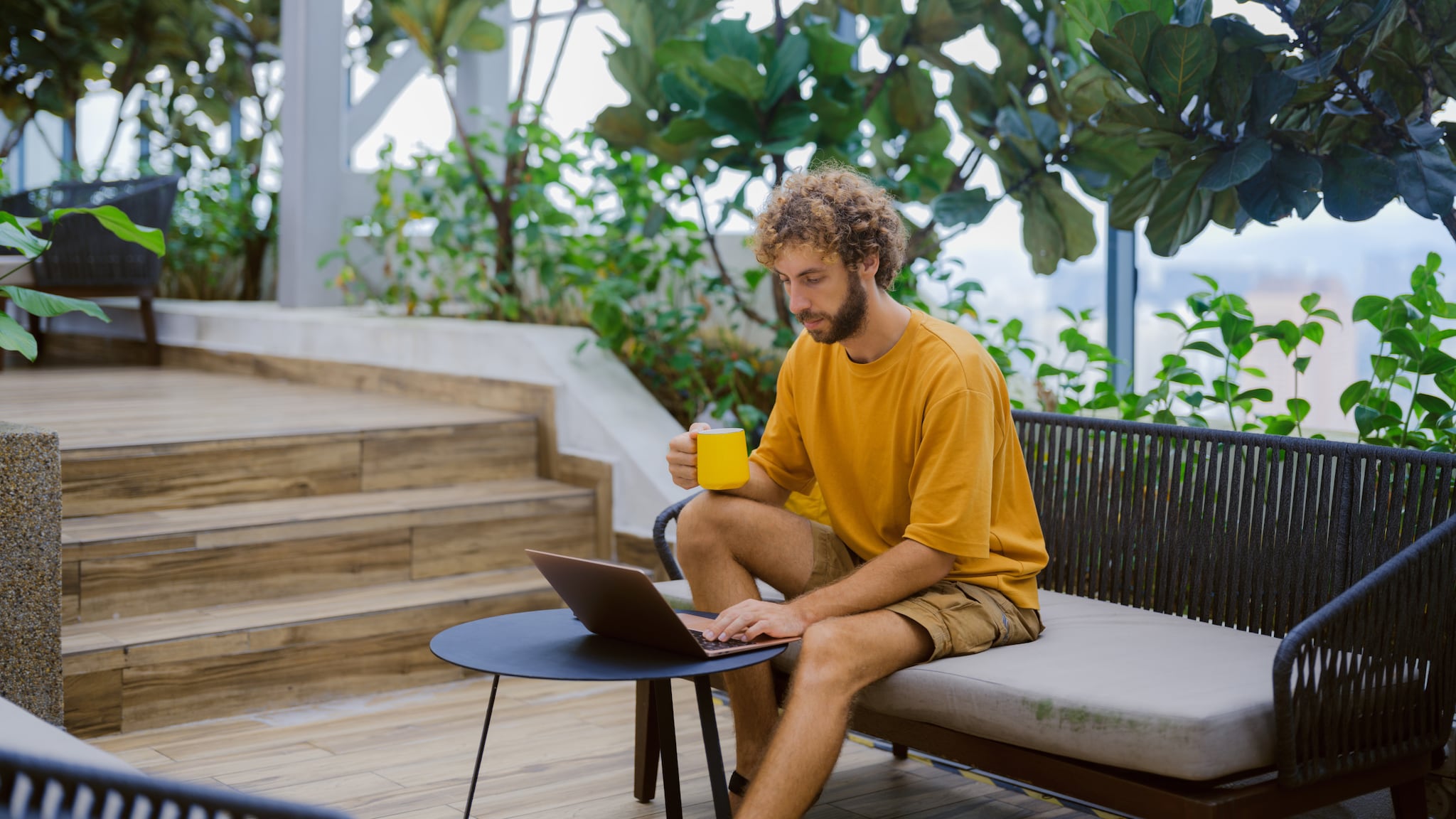 amex-sbs-vorteile-flexible-arbeitszeit-mann-laptop Ein Mann auf einer Terrasse tippt mit einer Hand auf einem aufgeklappten Laptop auf dem Tisch vor ihm, in der anderen Hand hält er eine Tasse.