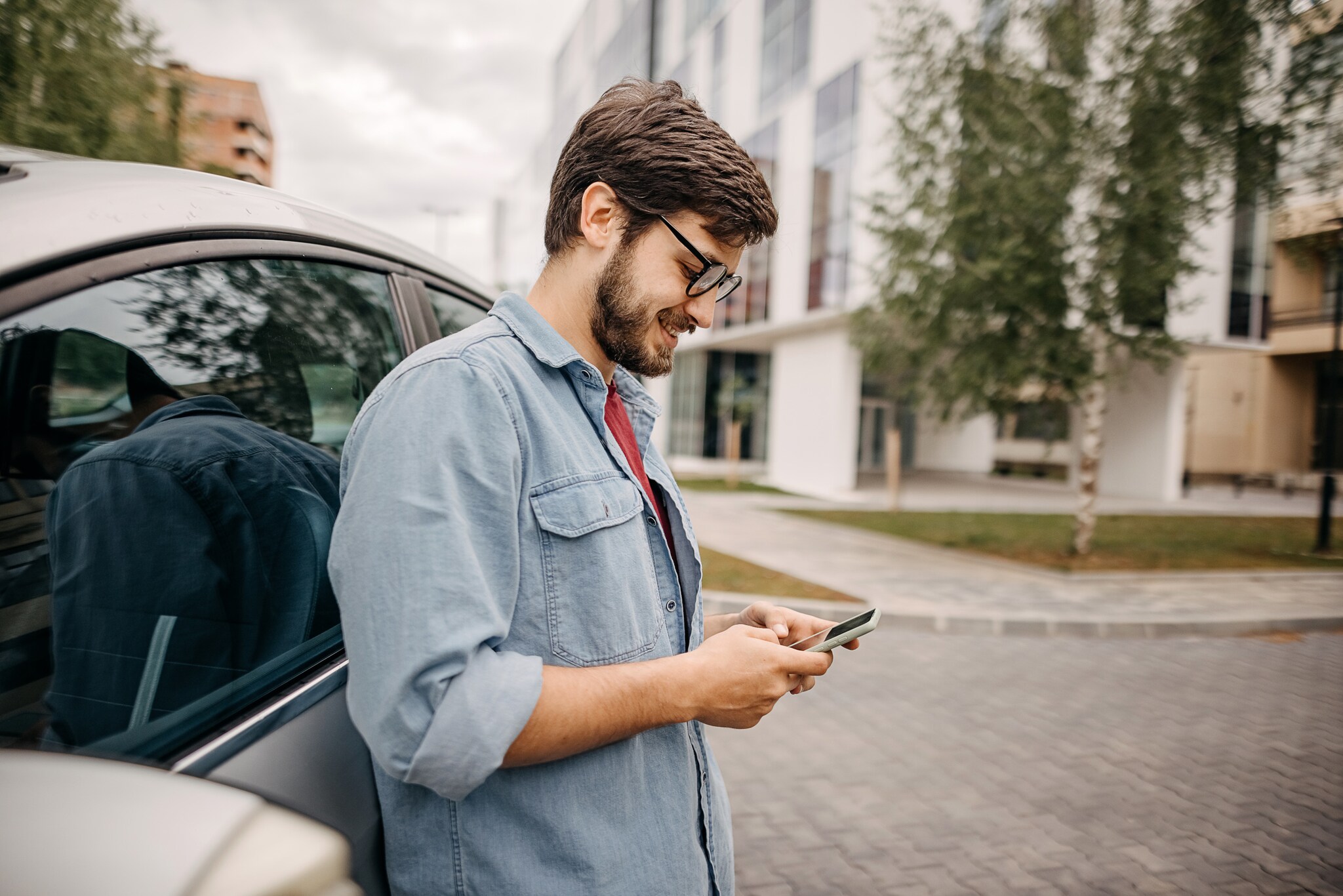 Mann, der an ein Auto lehnt und dabei ins Handy schaut.