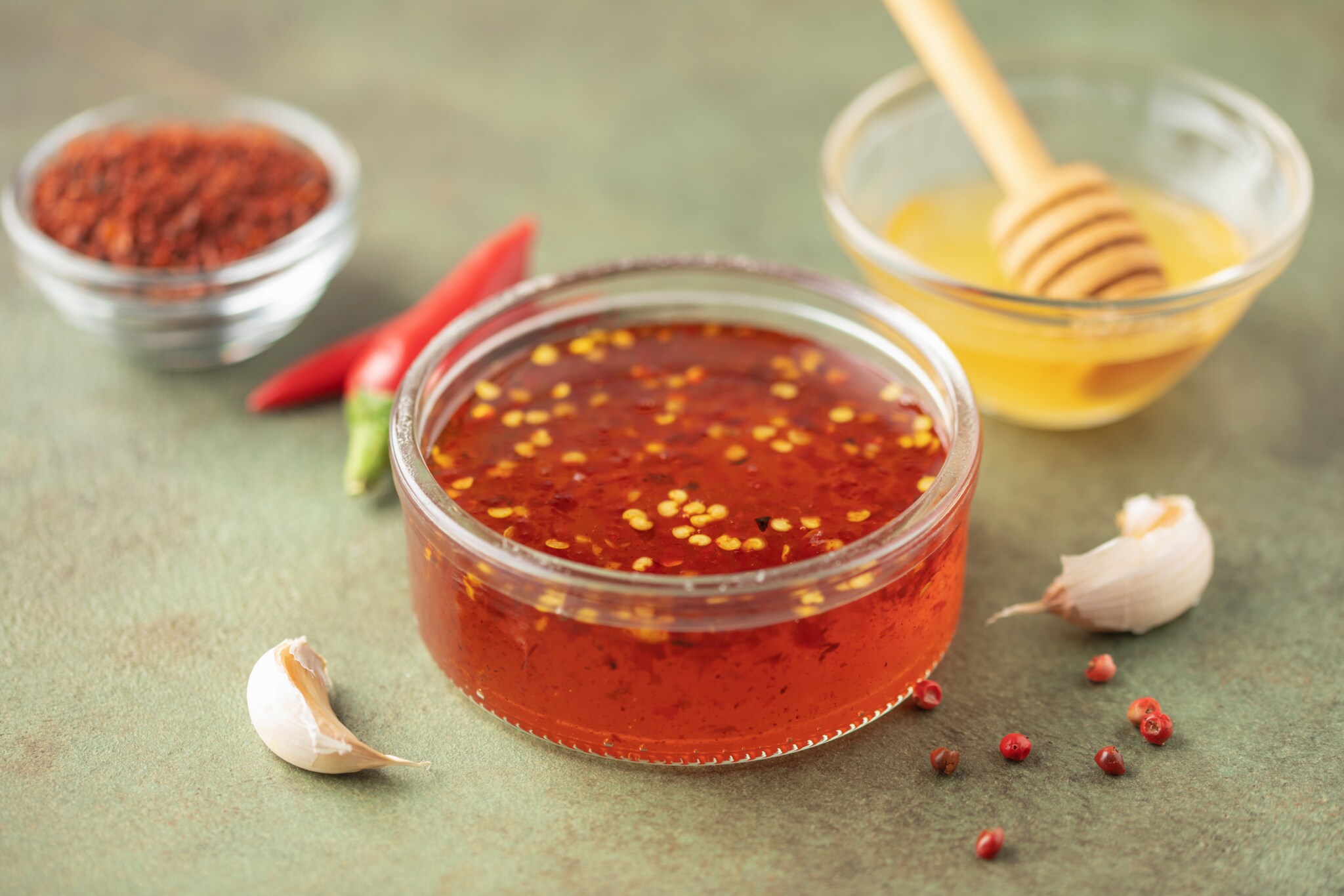Eine Schale mit Hot Honey, einem mit Chili gewürzten Honig steht auf einer grünlichen Unterlage, dahinter je eine Schale mit Chilipulver und reinem Honig.