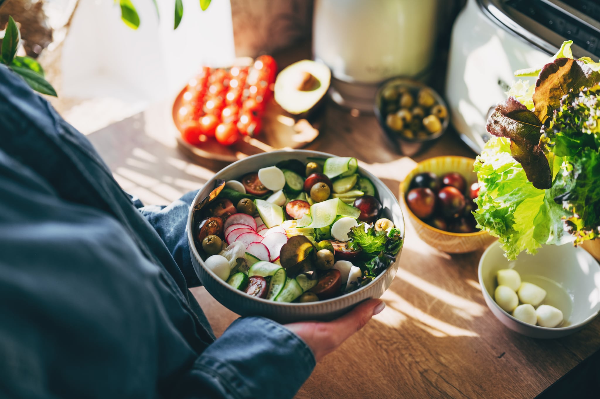 Eine Frau hält eine Bowl mit gesunden Zutaten in den Händen, im Hintergrund stehen Schalen mit frischem Gemüse.