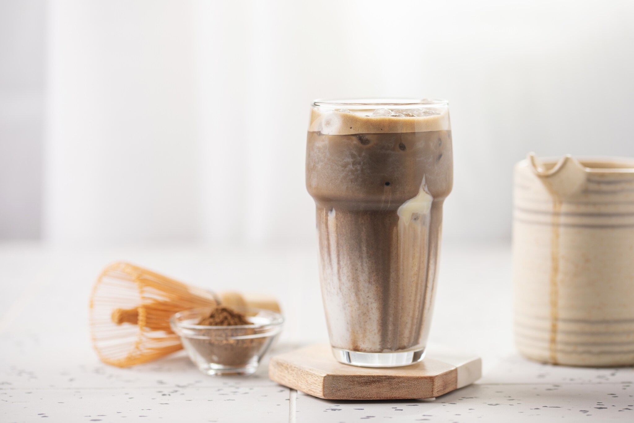 Ein erfrischender kalter Eis-Hojicha-Latte in einem hohen Glas steht neben einem Chasen-Bambusbesen, einer Kanne und einer kleinen Schale mit Hojicha-Pulver.