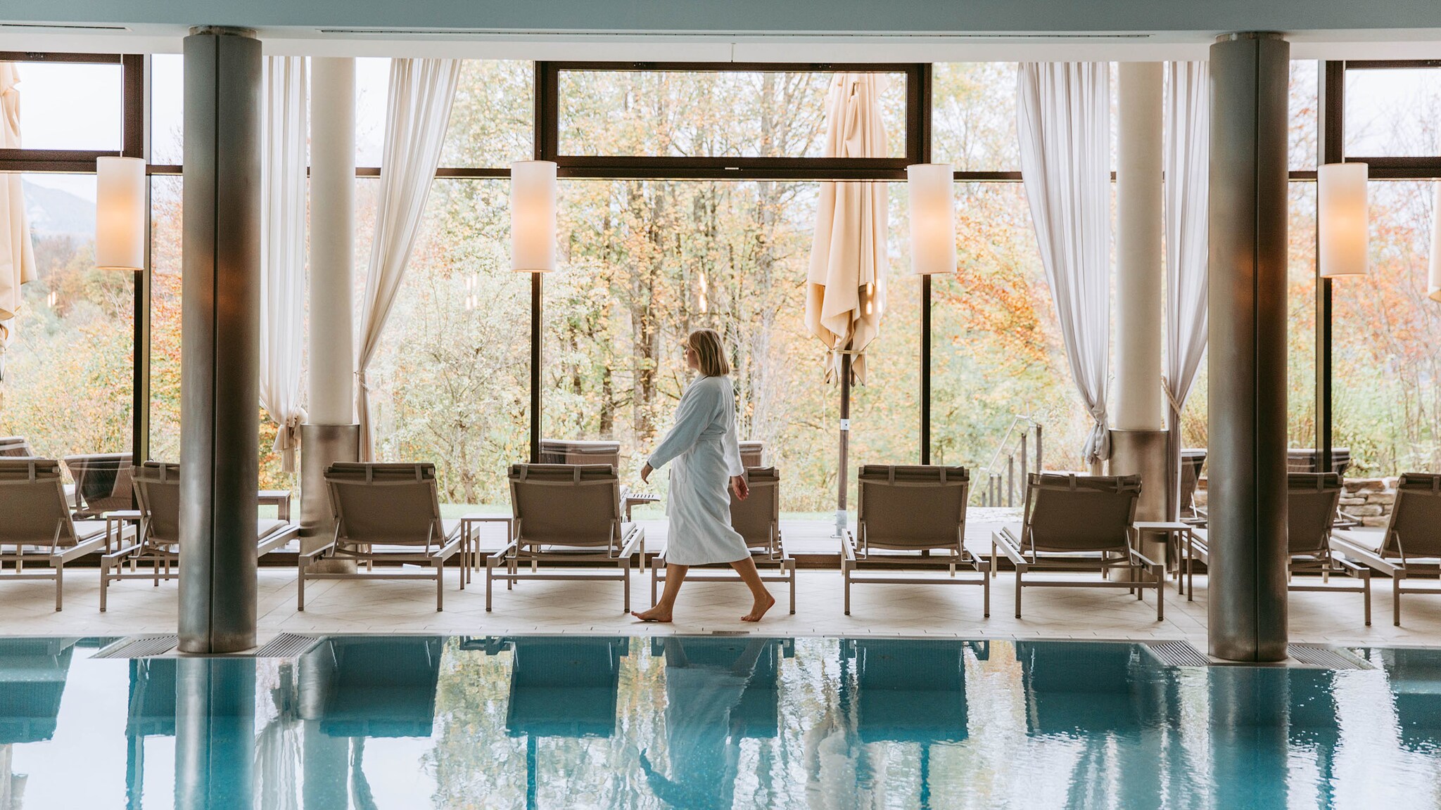 Eine Frau im weißen Bademantel geht an einem Indoorpool entlang, vorbei an Liegestühlen vor einem Panoramafenster mit Blick in eine herbstliche Waldlandschaft. 