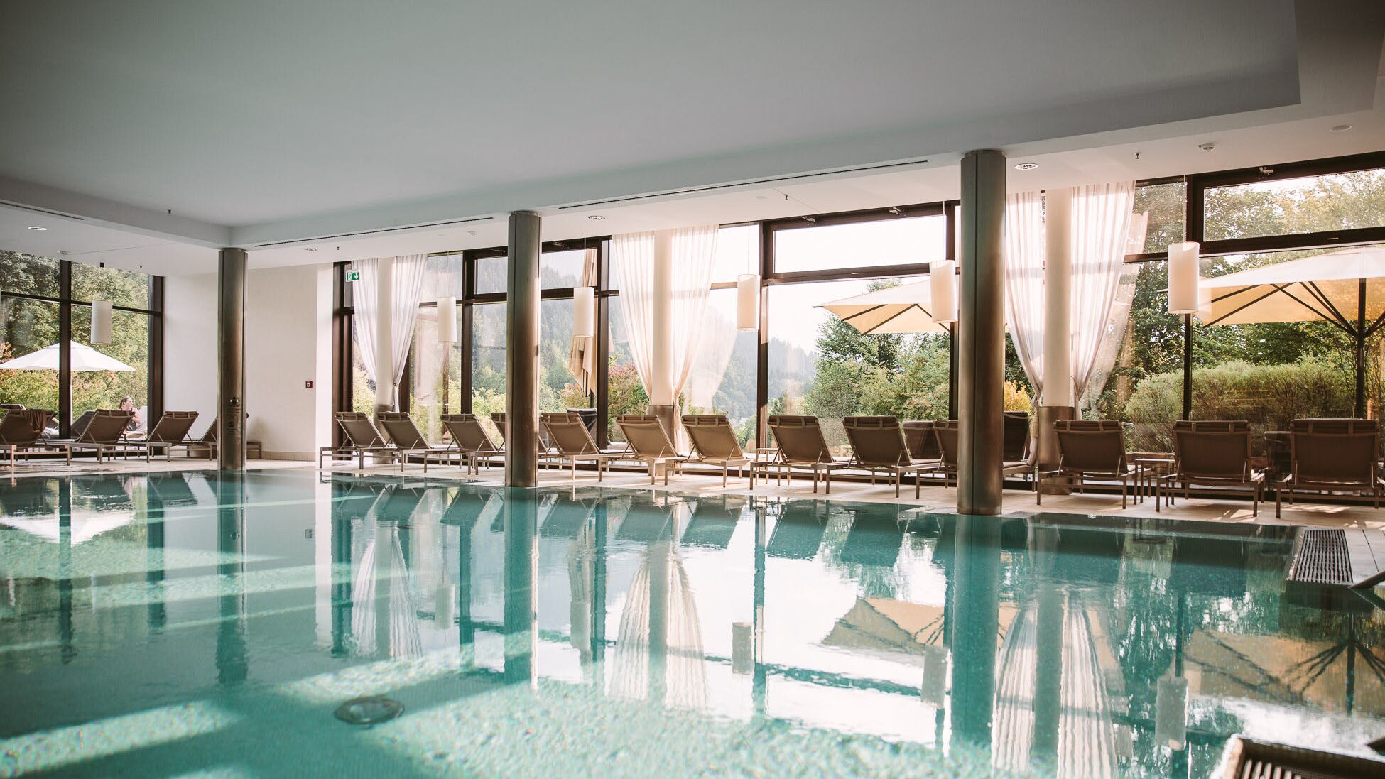 Indoorpool, umgeben von Liegen und einer Fensterfront mit Blick in eine bergige Waldlandschaft.