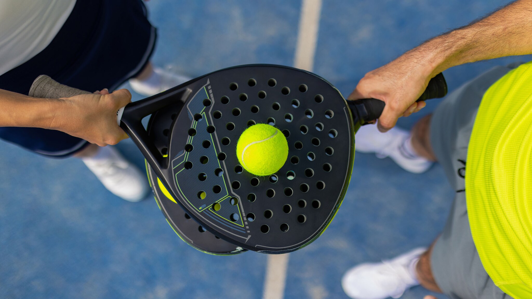 Zwei Padelschläger werden von zwei Personen übereinander gehalten, darauf liegt ein gelber Tennisball.
