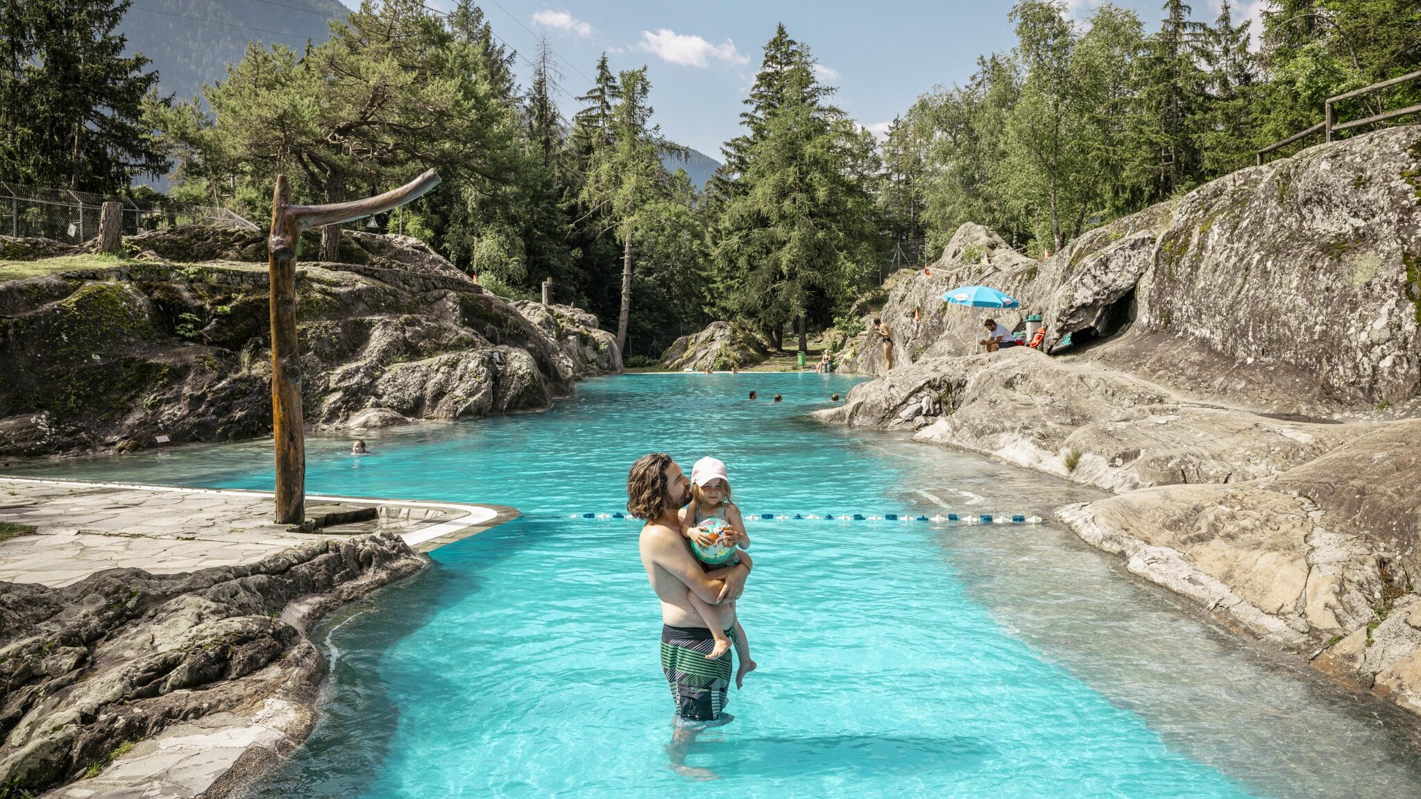 Ein Mann steht im Naturbad Les Marécottes und hält ein Kind auf dem Arm.