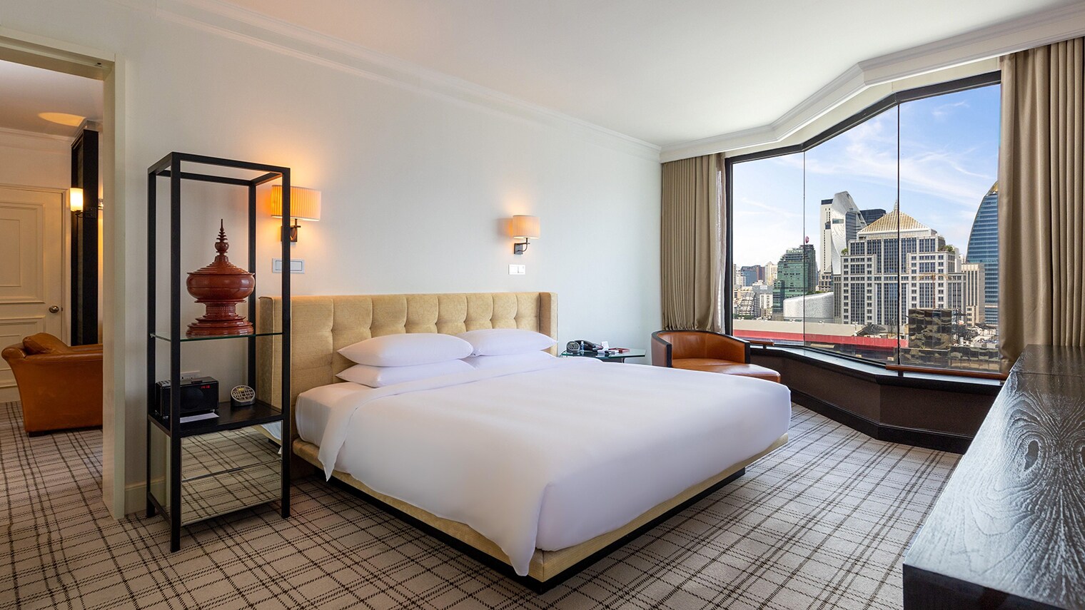In einer Hotelsuite steht ein großes Bett vor einem großen Fenster, durch das man die Skyline einer Stadt sehen kann.