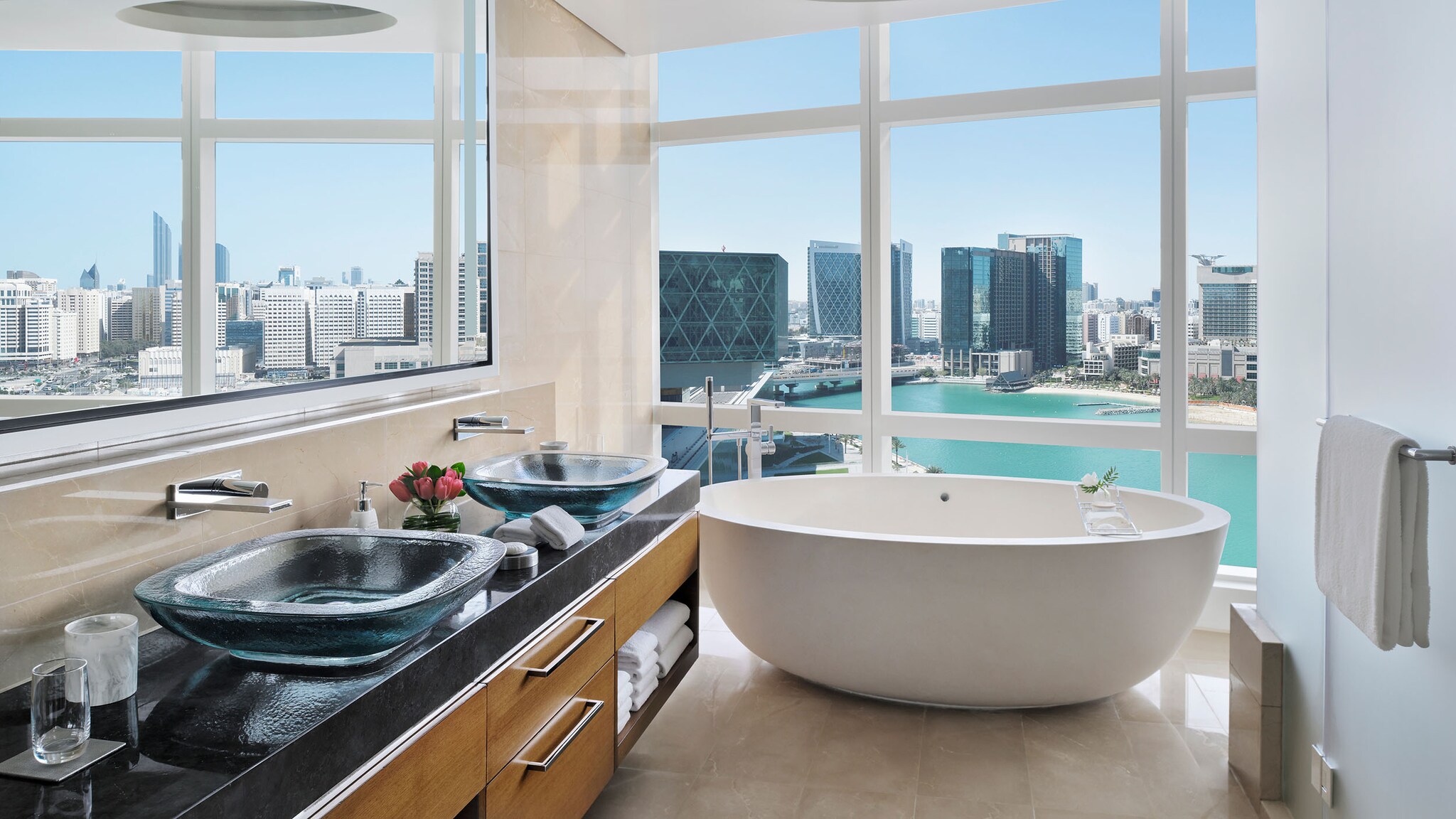 In einem Badezimmer steht ein Waschtisch und eine Badewanne vor einem bodentiefen Fenster mit Blick auf die Skyline einer Stadt.