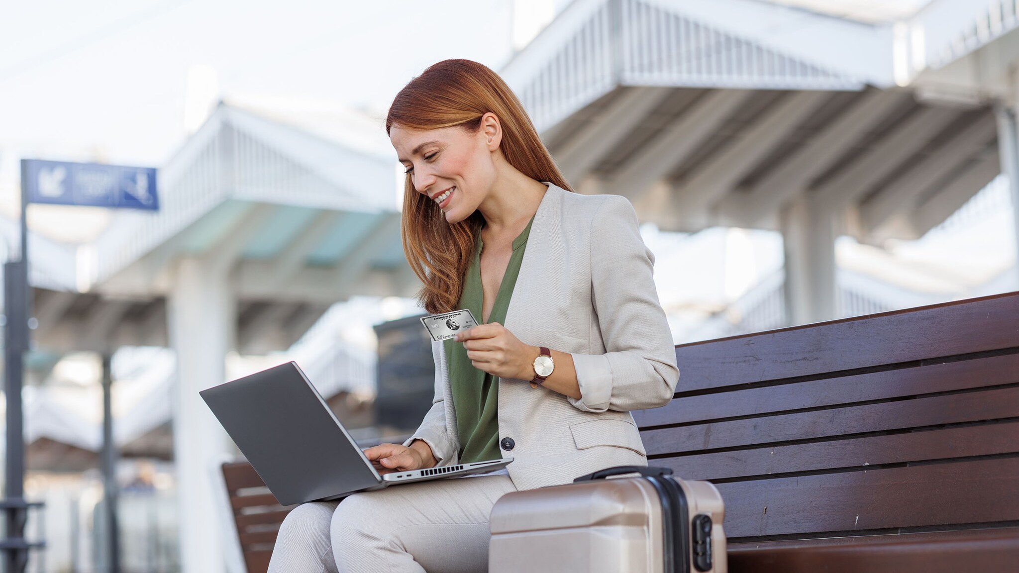 Eine Frau sitzt auf einer Bank an einem Bahnhof, hat ihren Laptop auf dem Schoß und eine Kreditkarte in der Hand.