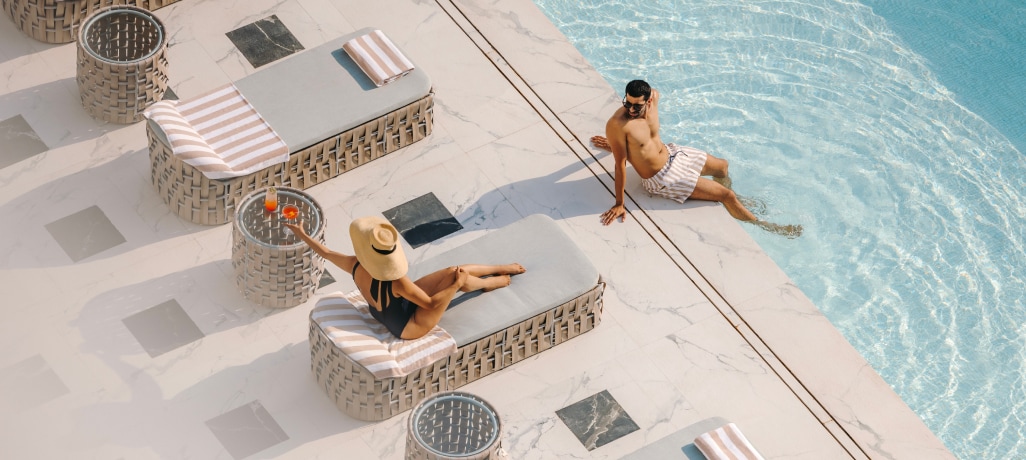ALL Accor Bonusprogramm Eine Frau mit Sonnenhut liegt auf einer Liege an einem Pool, an dessen Rand ein Mann sitzt bei Sonnenschein.