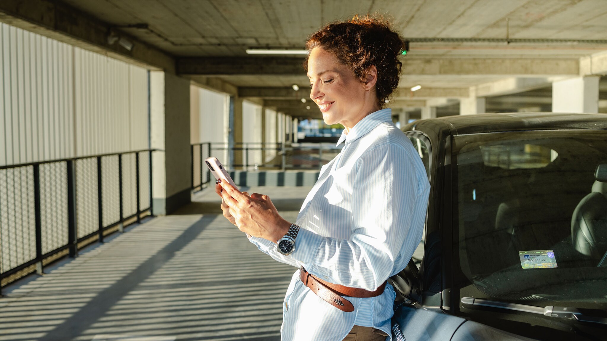 Eine Frau steht in einer Tiefgarage an einem Auto und schaut auf ihr Smartphone.