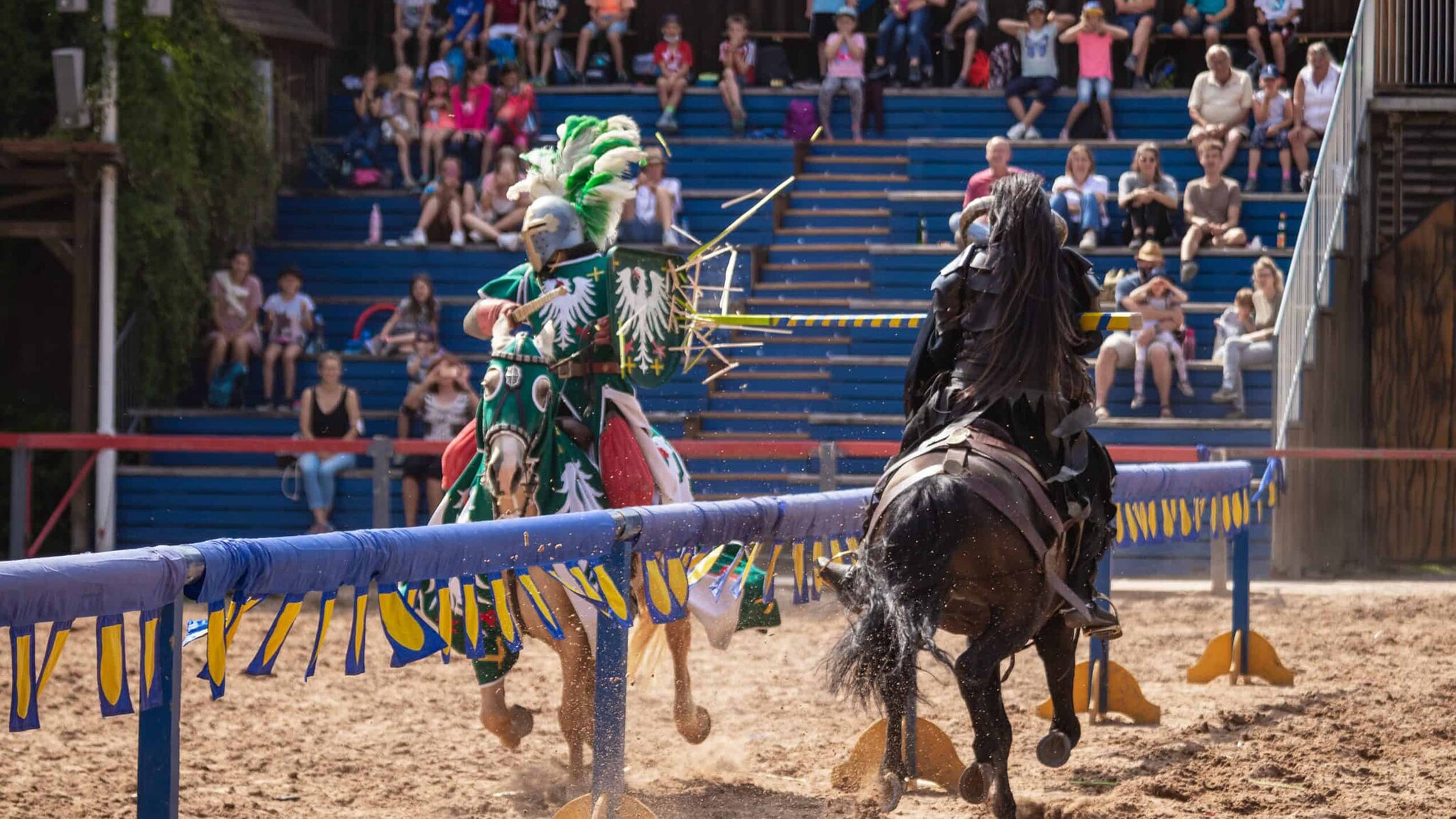 Auf einem Festplatz reiten zwei Persone in Ritterrüstung und Lanze aufeinander zu.
