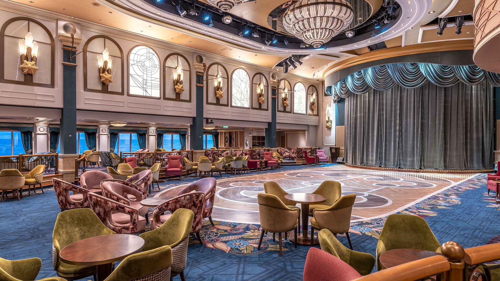 Im eleganten Queens Room stehen Loungemöbel vor einer Bühne, an der Decke hängen große Kronleuchter.