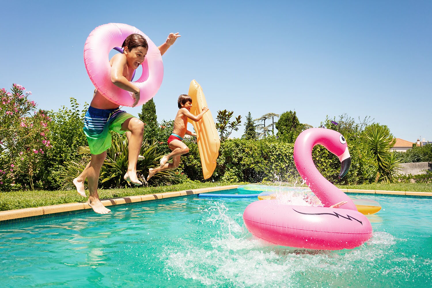 Zwei Kinder springen mit Schwimmreifen in eine Pool mit Schwimmtier bei Sonnenschein.