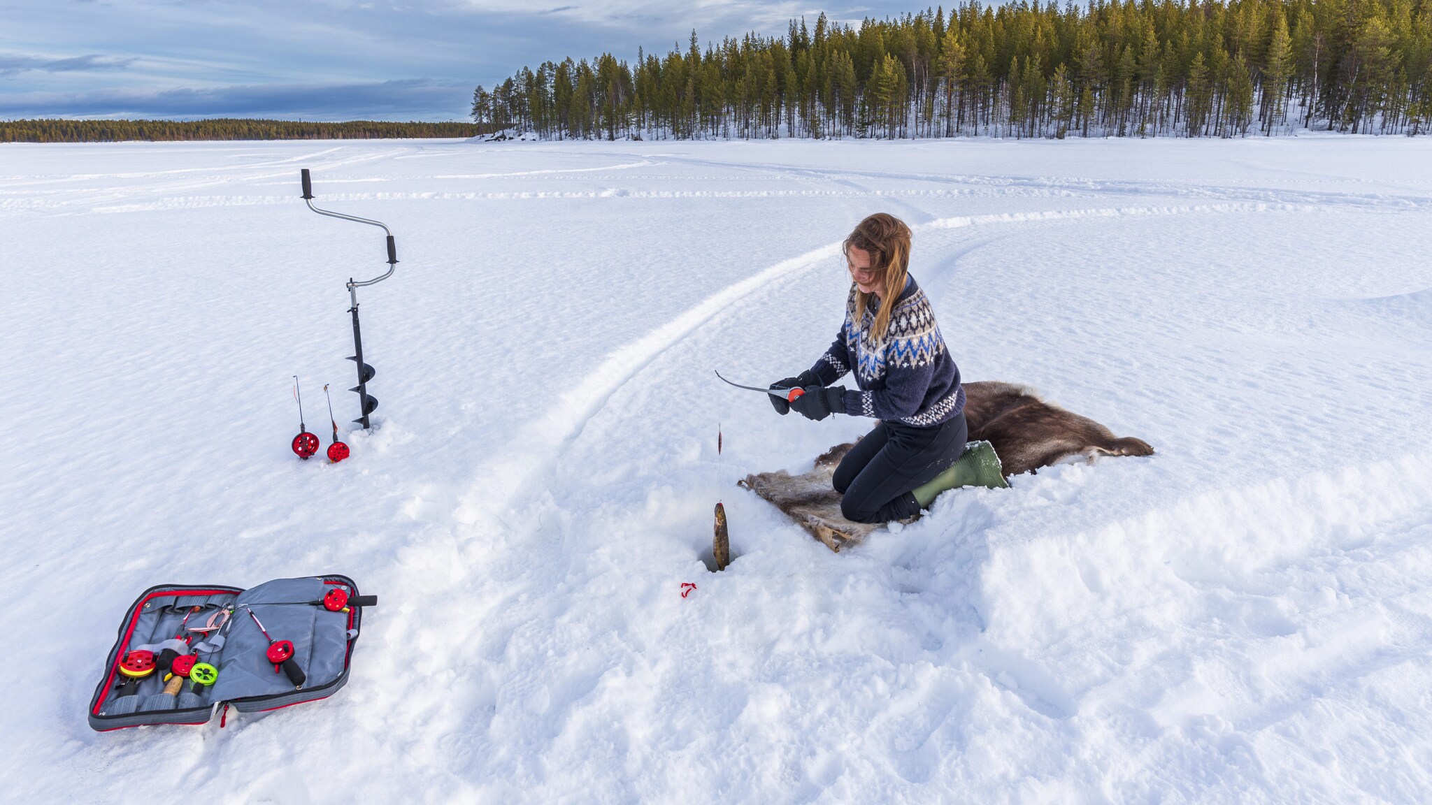 Eine Frau kniet auf einem Fell in einer schneebedeckten Landschaft und angelt einen Fisch aus einem Loch im Eis.