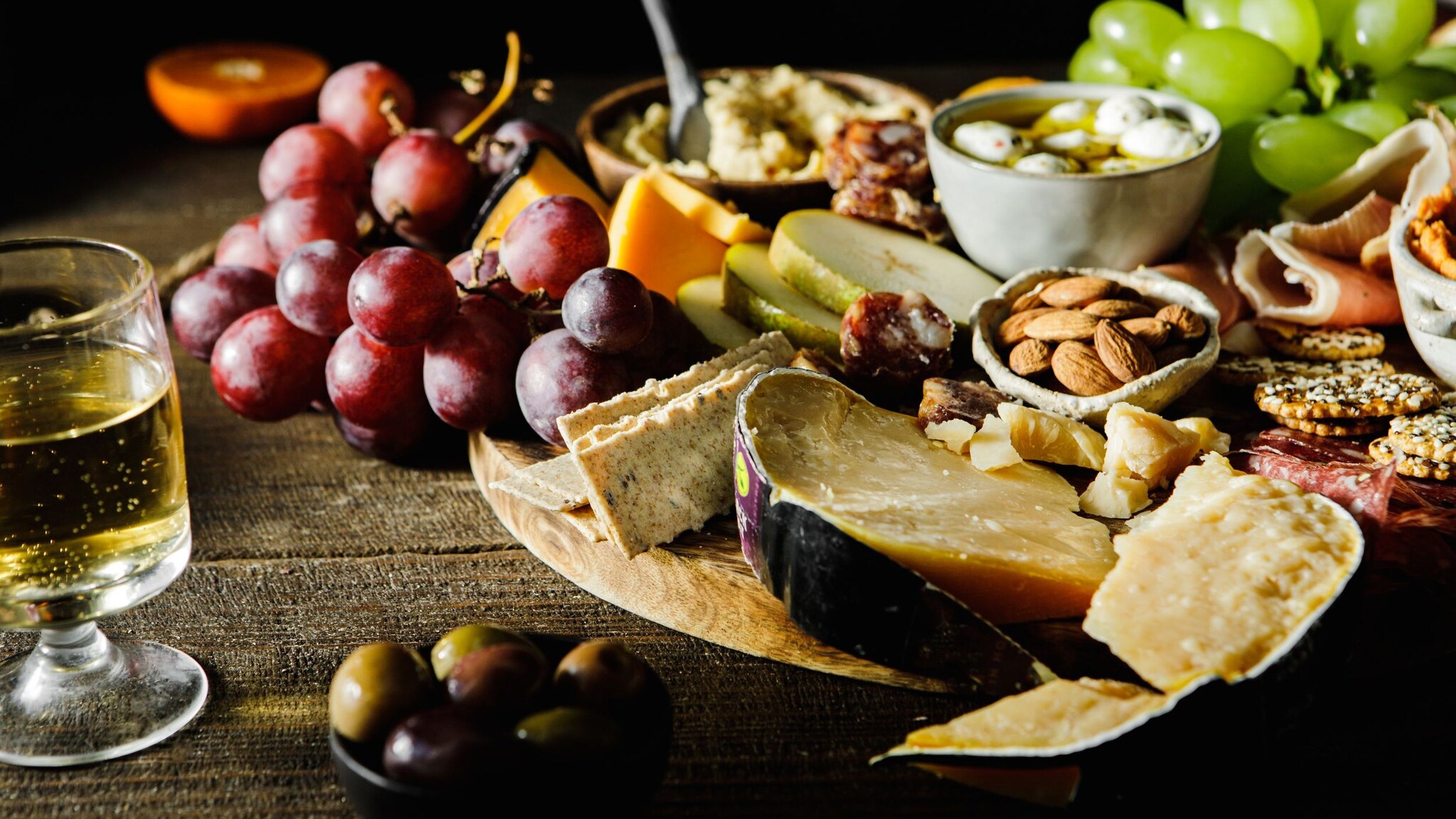 Glas Weißwein auf einem Holztisch neben einem Brett mit verschiedenen Snacks. 