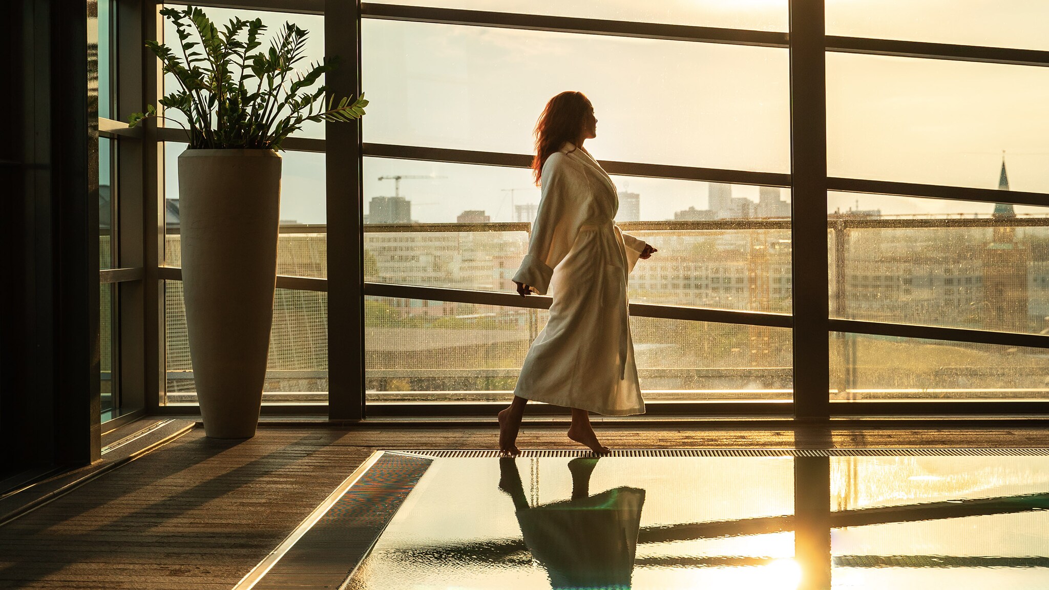 Eine Frau im weißen Bademantel läuft am Rande eines Indoorpools vor einem Panoramafenster mit Blick auf die Berliner Skyline im Sonnenlicht.