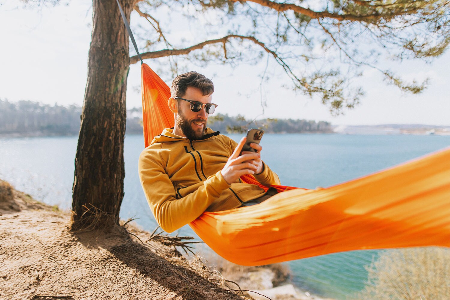 Ein Mann mit Sonnenbrille liegt in einer Hängematte an einer Meeresküste und blickt auf ein Smartphone in seinen Händen.