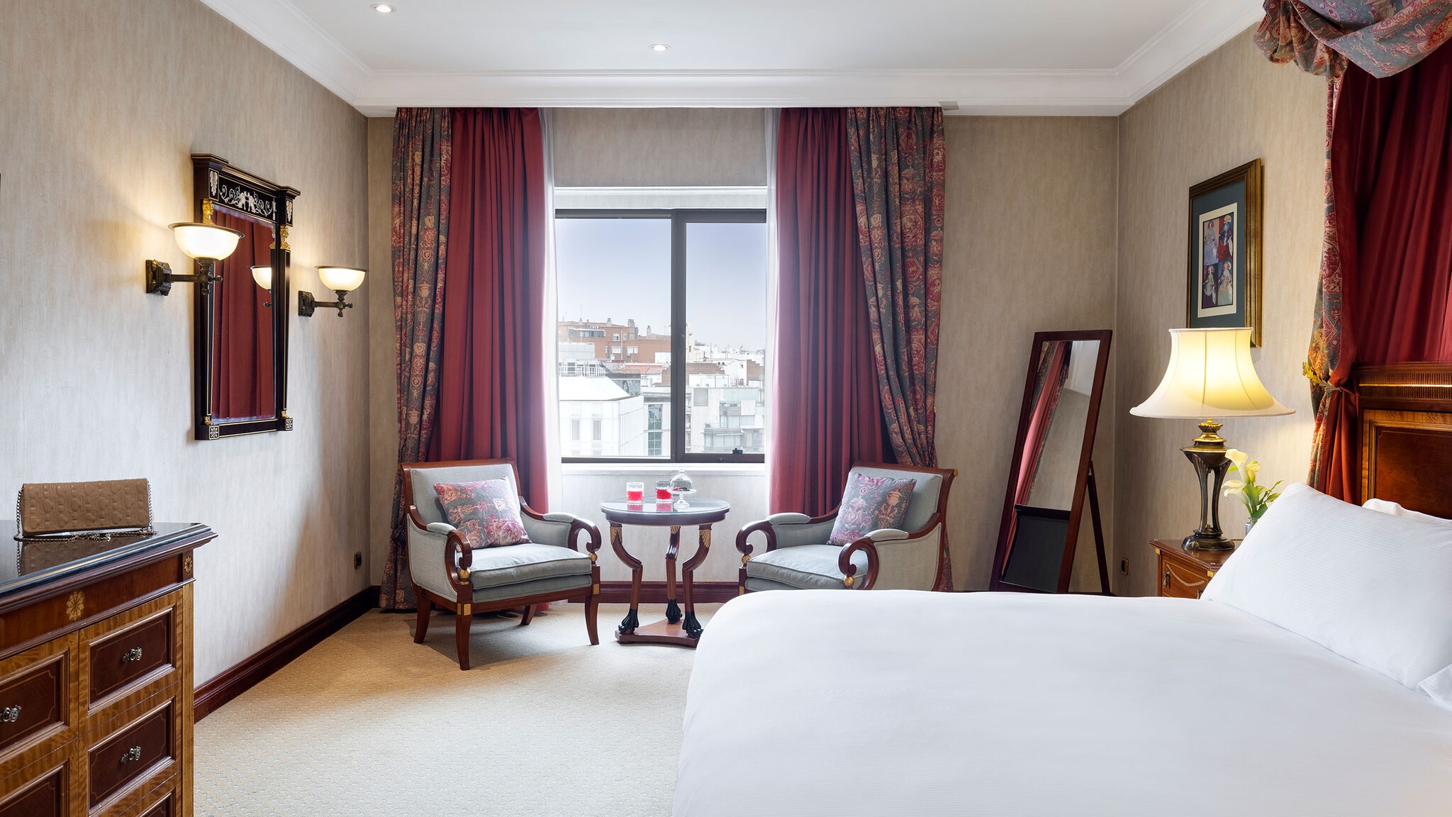 In einem Hotelzimmer steht ein Bett und weitere Möbel, die Fenster haben rote Vorhänge.