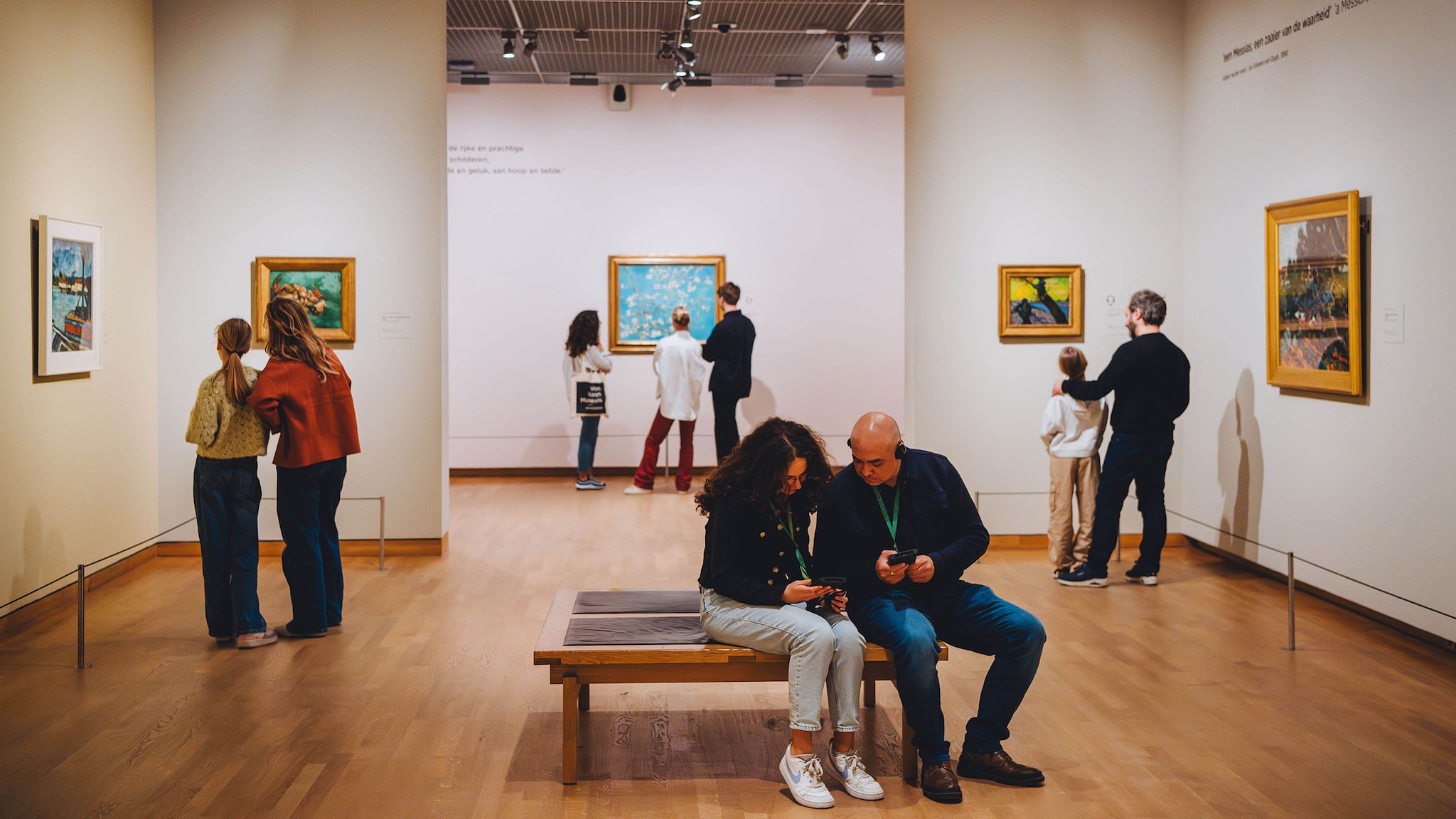 In einem Museum hängen Gemälde des Malers Van Gogh an den Wänden, Besucher stehen davor oder sitzen auf Bänken.
