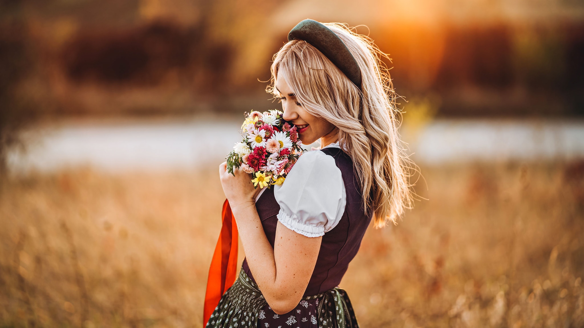 Eine Frau trägt ein Dirndl, steht auf einer Wiese und riecht an einem Blumenstrauß.