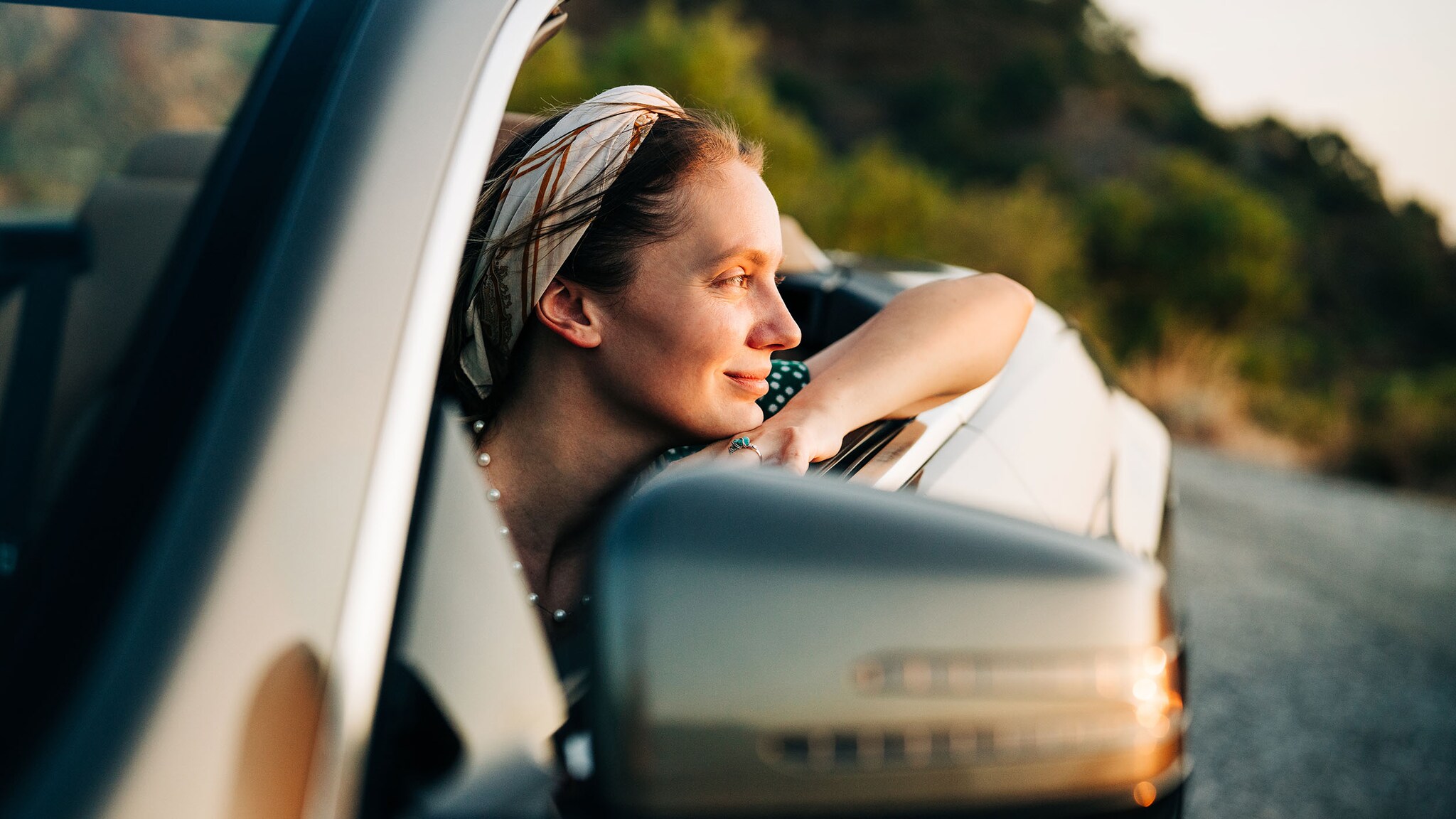Porträt einer zufriedenen lächelnden Frau, die mit aufgestütztem Arm vom Sitz eines Cabriolets blickt auf einer Landstraße im weichen Sonnenlicht.
