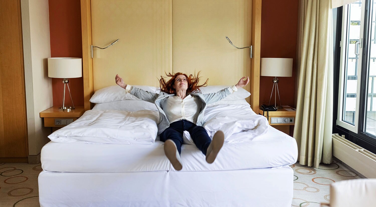 Eine lachende Frau liegt in Straßenkleidung rücklings mit ausgestreckten Armen auf einem Hotelbett.