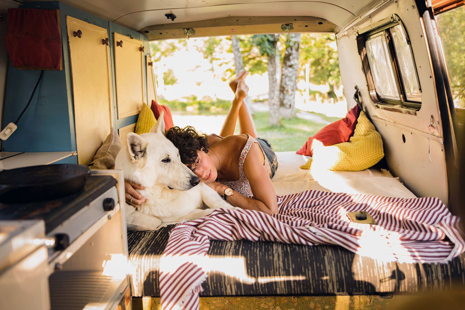 Eine Frau in Sommerkleidung liegt auf dem Bauch mit angewinkelten Beinen in einem Campervan mit geöffneter Tür und kuschelt mit ihrem Hund in einem Waldgebiet.