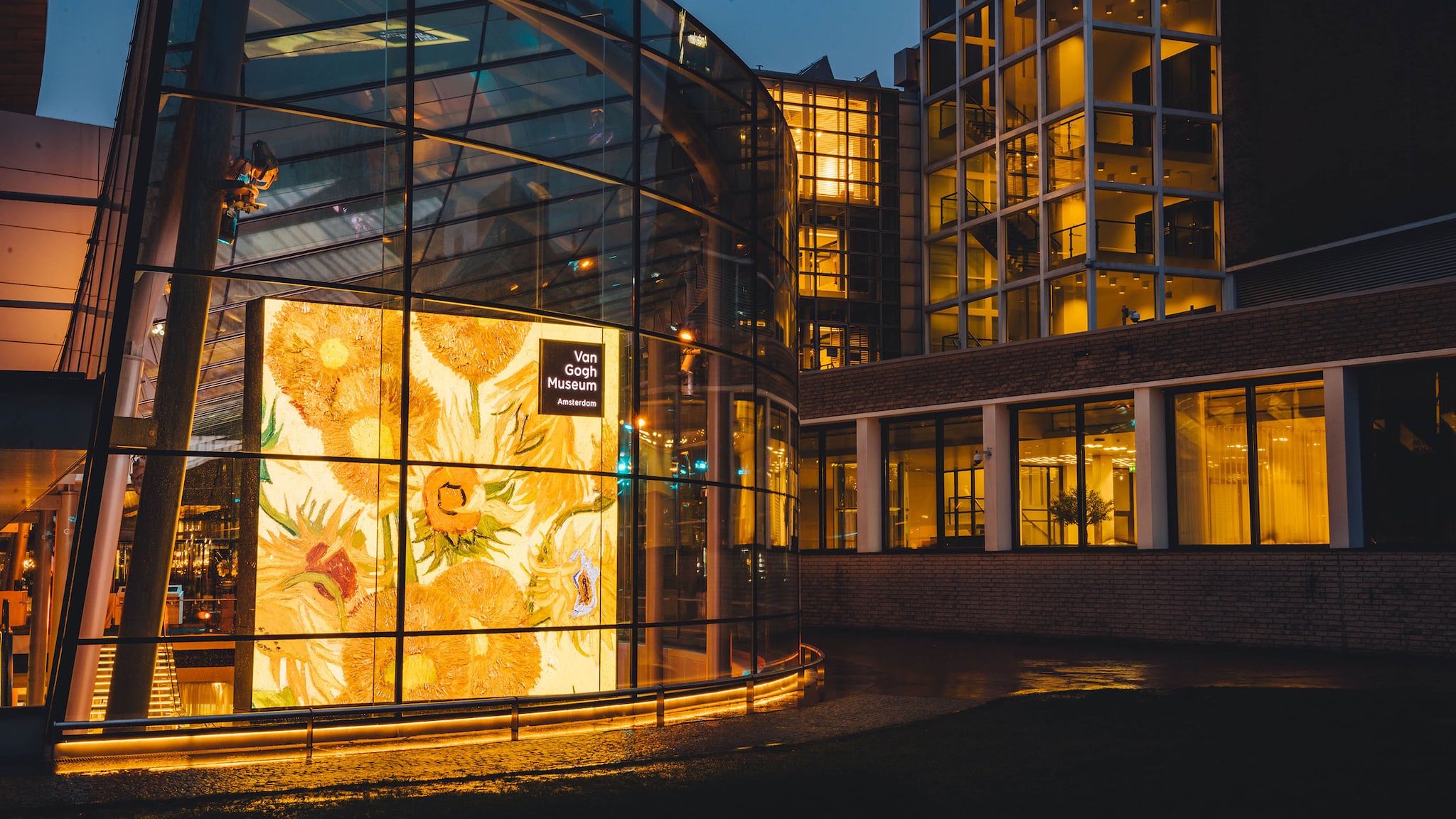 Das Außengebäude des Van Gogh Museums hat eine große, beleuchtete Glasfront, in der ein großes Bild mit Sonnenblumen steht. 