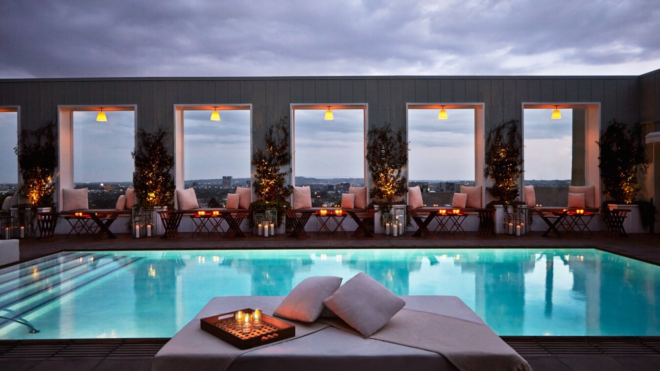 An einem Hotelpool stehen kleine gedeckte Tische mit Kerzen und gemütlichen Stühlen.