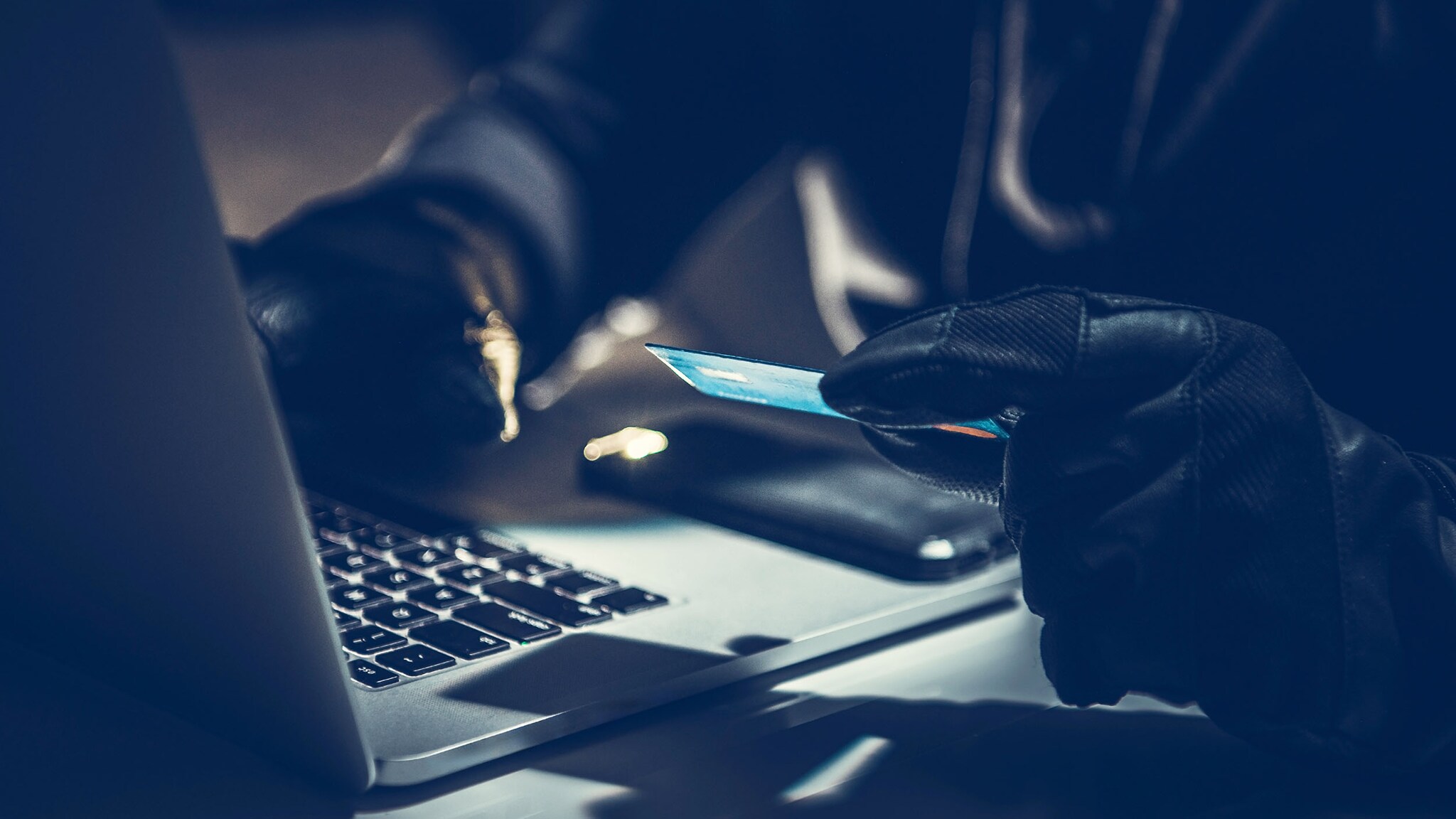 Nahaufnahme von zwei Händen in schwarzen Lederhandschuhen an einem Laptop, eine Hand hält eine Kreditkarte im schummrigen Licht.