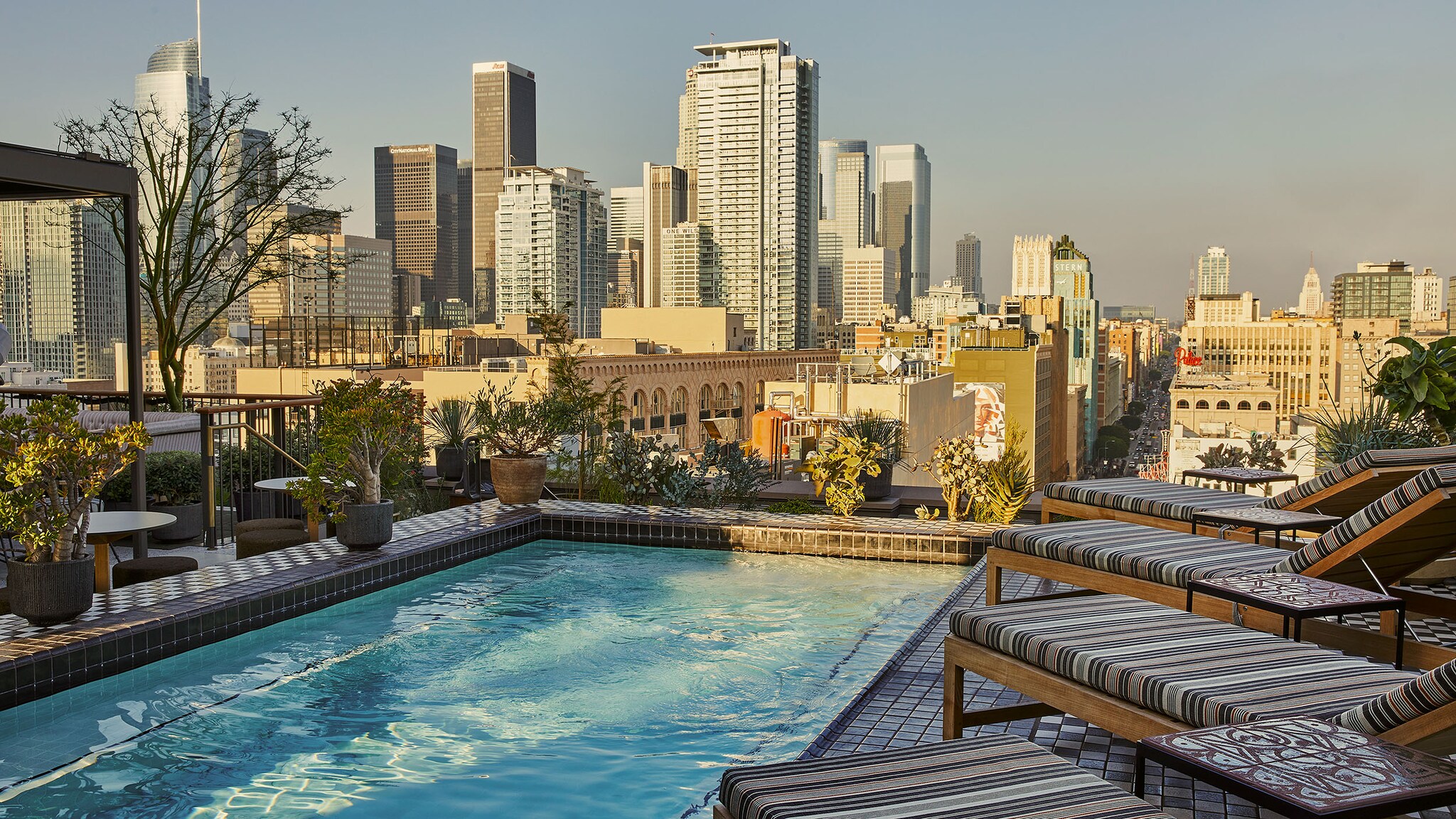 Auf der Dachterrasse eins Hotels ist ein Pool mit Sonnenliegen, im Hintergrund sieht man die Skyline von Los Angeles.