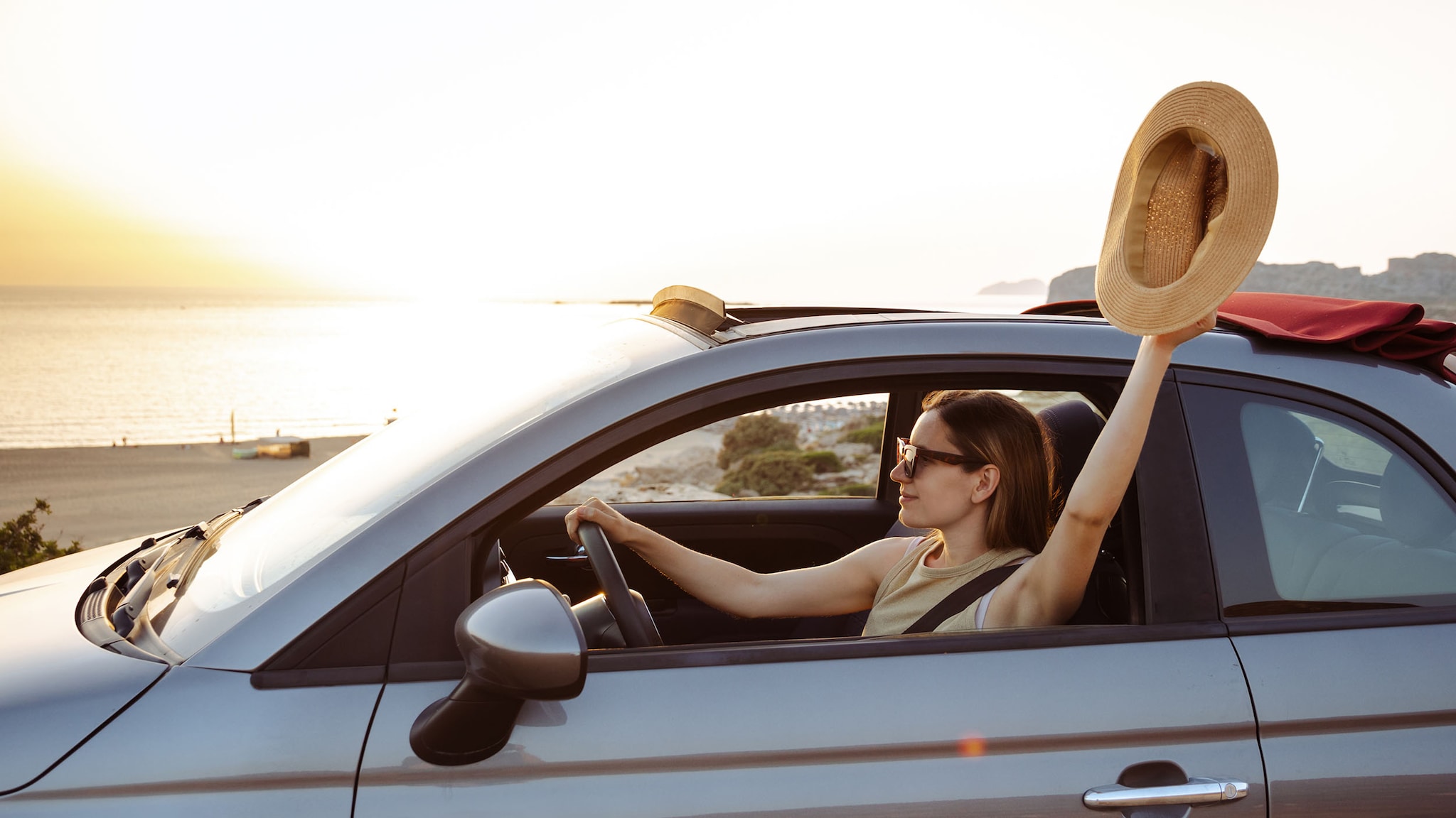 Eine Frau fährt in einem Auto und hält ihren Sonnenhut aus dem Fenster.