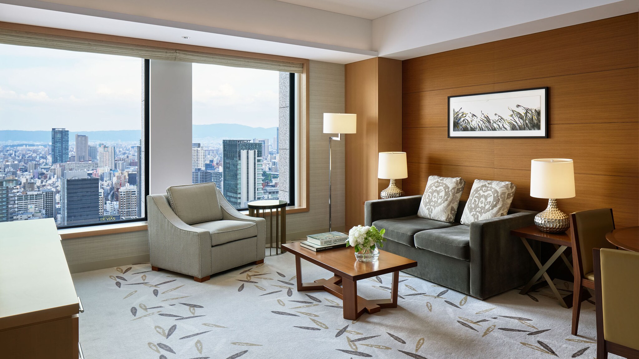In einem Hotelzimmer steht ein Sofa und ein Sessel vor einem Fenster, von dem man die Skyline einer Stadt sieht.
